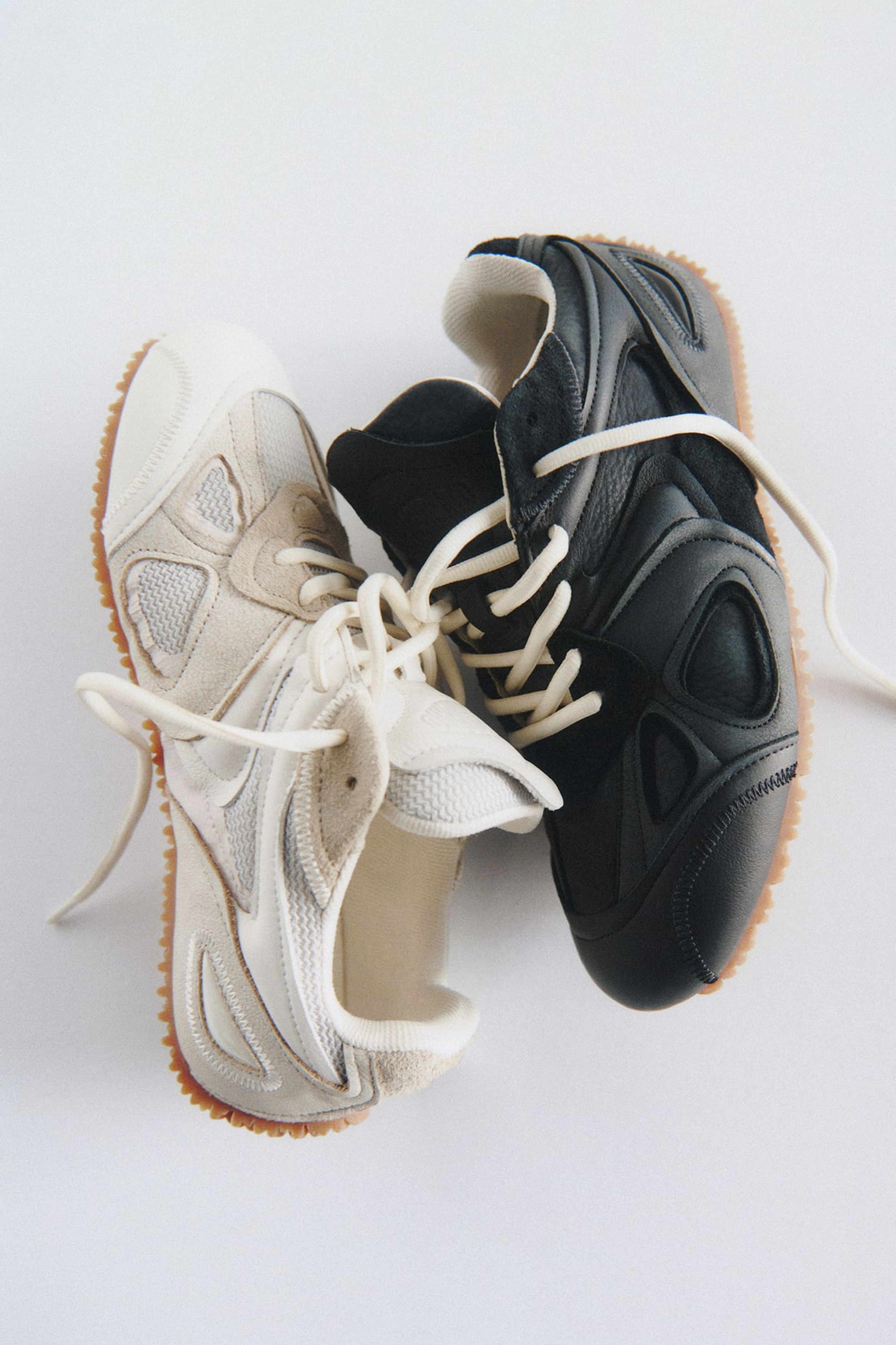 Two sneakers, one white and one black, are intertwined with their laces on a white surface, featuring gum soles and mesh detailing.