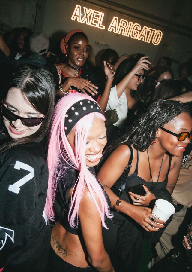 People dancing joyfully at a party under neon "AXEL ARIGATO" sign. A woman with pink hair and others smile and enjoy the lively atmosphere.