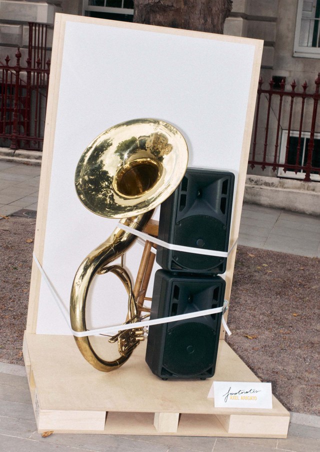 A sousaphone is strapped to two stacked speakers on a wooden platform outdoors, with a small sign nearby.