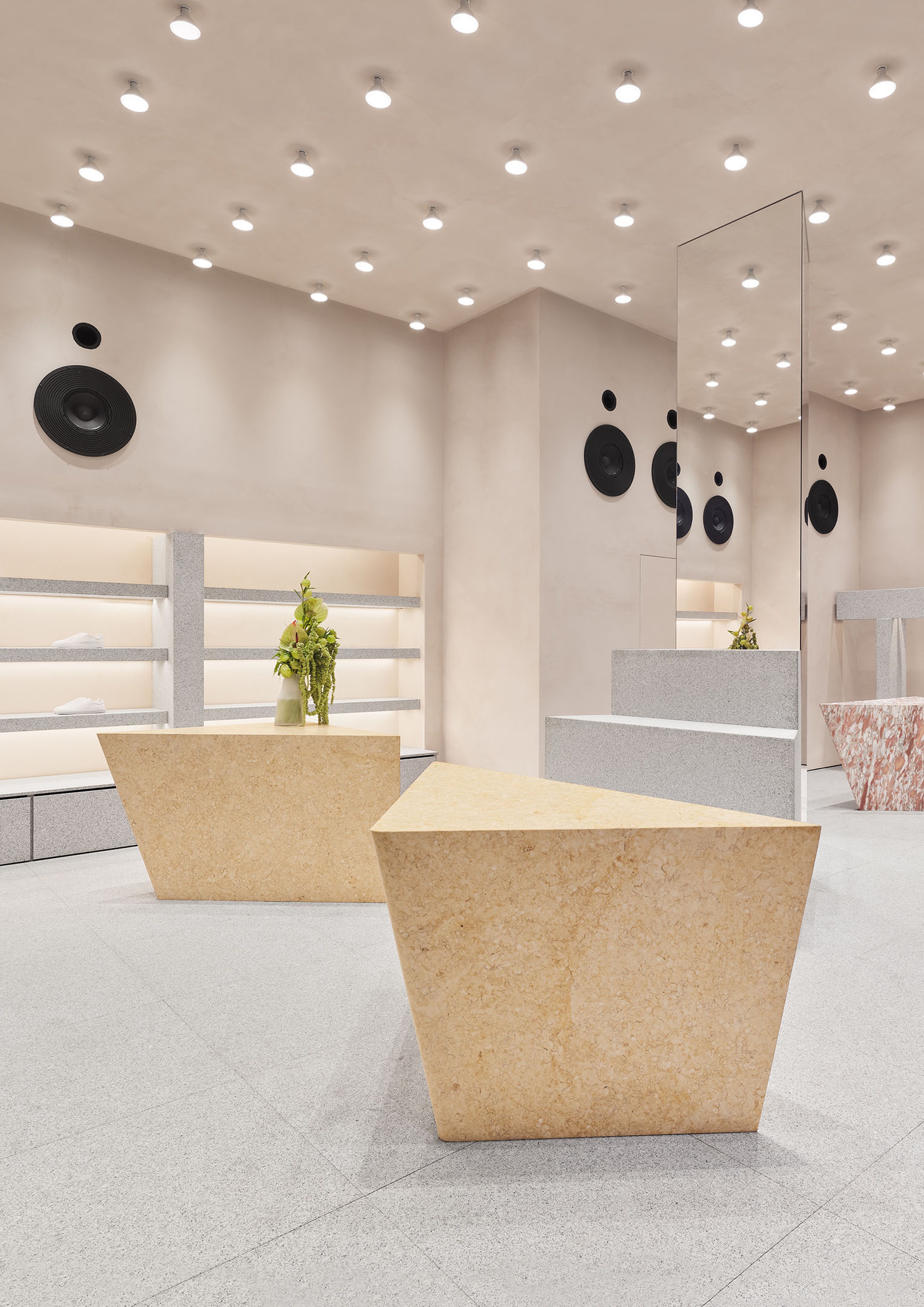 Modern retail space with geometric stone tables, mirrored wall, and shelves. Ceiling has recessed lighting and black circular speakers.