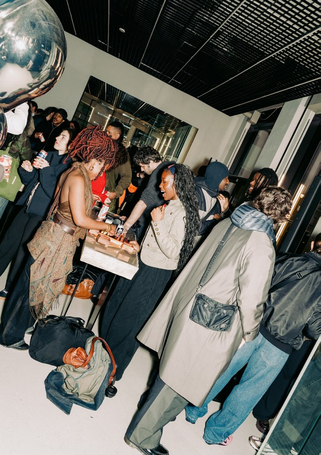 A lively indoor gathering with people socializing and enjoying food. Some are standing near a table, while others mingle in the background.