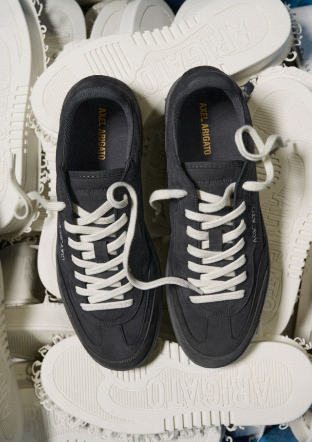 Black sneakers with white laces rest on a pile of white sneaker soles. "Axel Arigato" is printed on the insoles and side of the shoes.