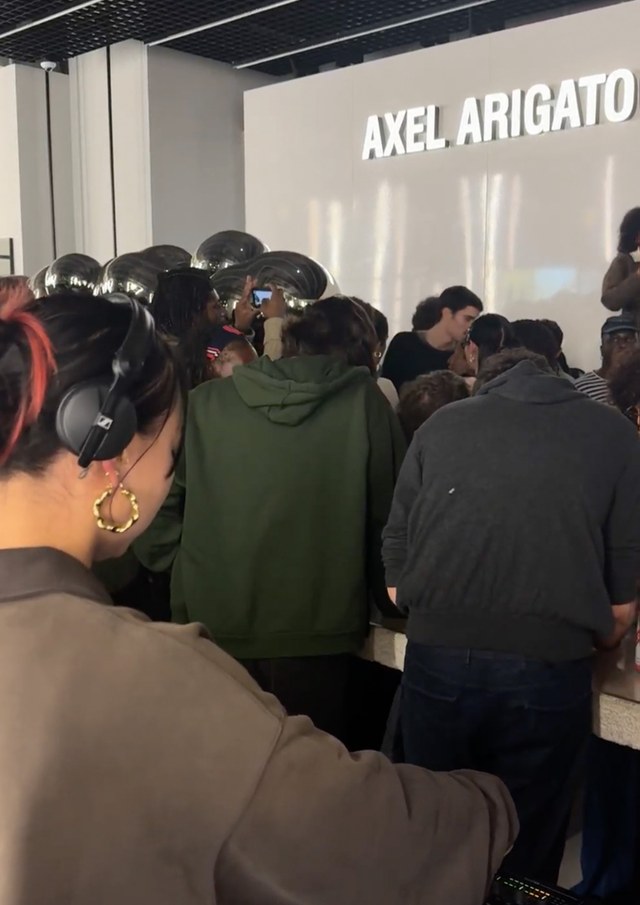 A DJ with headphones stands at a console, facing a crowd gathered near a wall with "AXEL ARIGATO" written on it.