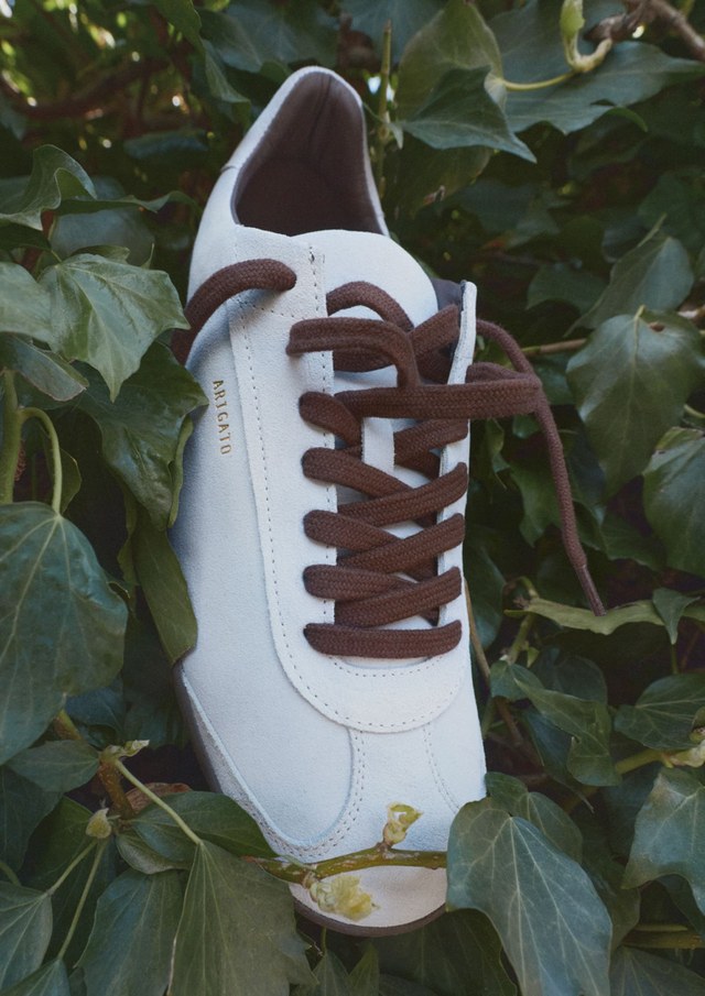 White sneaker with brown laces hanging in green foliage, showing a brand name on the side.