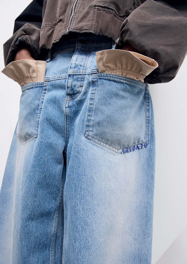 Person wearing high-waisted, loose-fitting blue jeans with large tan inside-out pockets and "ARIGATO" embroidered, paired with a dark cropped jacket.