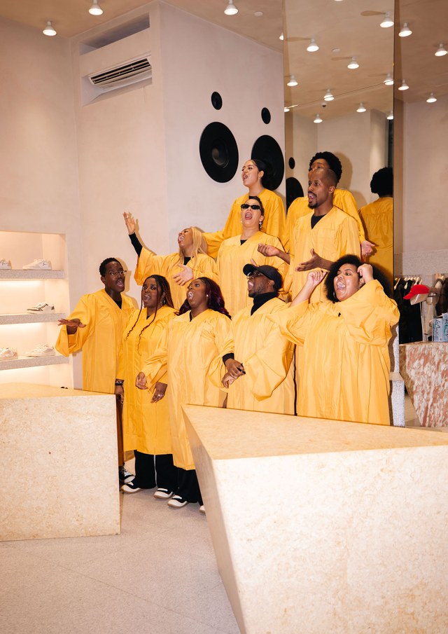 A choir wearing yellow robes sings joyfully in a modern room with geometric decor and shoes displayed on shelves.