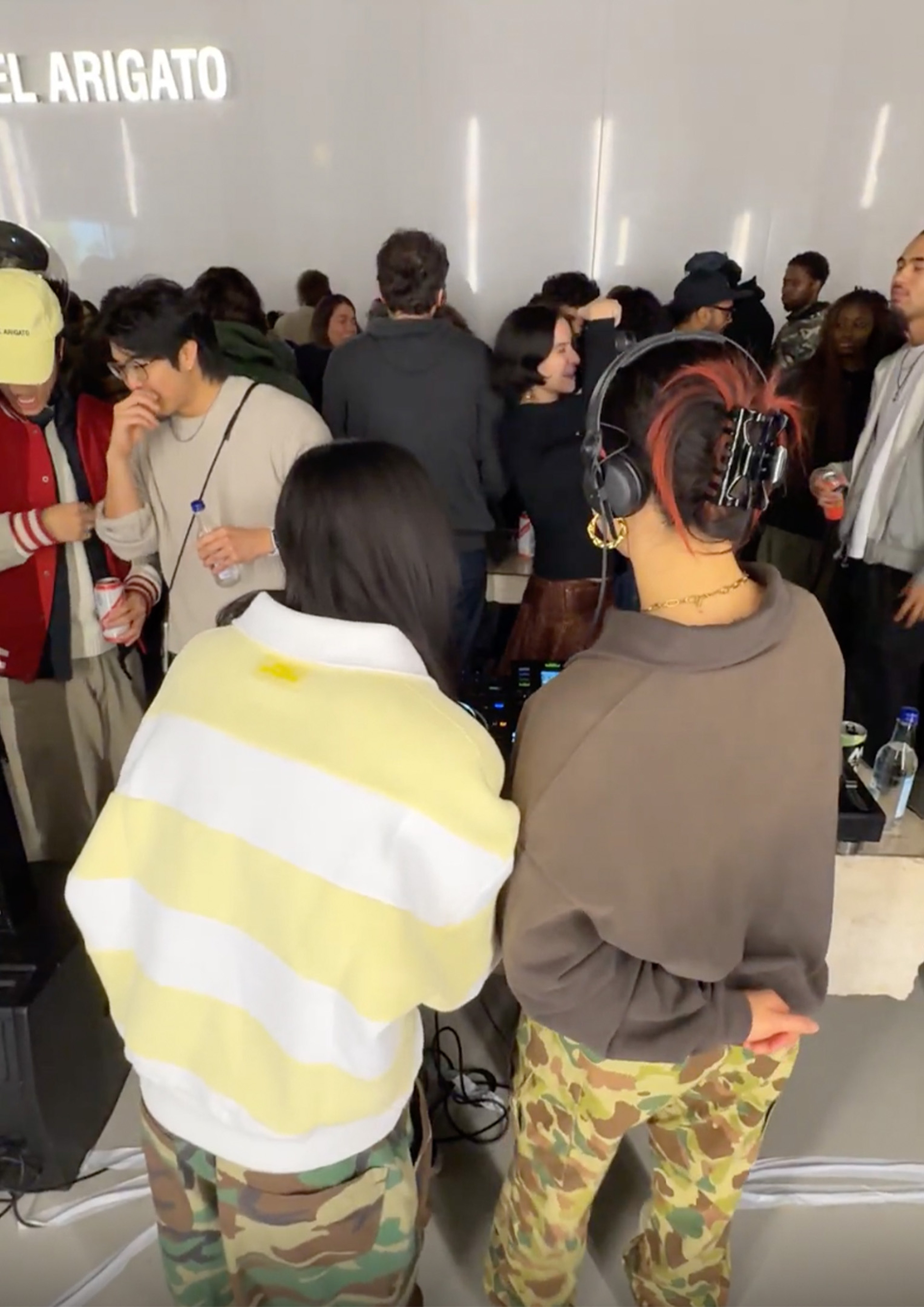 Two DJs in camouflage pants perform for a crowd in a lively, indoor setting. People are socializing and enjoying music in the background.