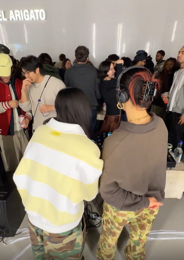 Two DJs in camouflage pants perform for a crowd in a lively, indoor setting. People are socializing and enjoying music in the background.
