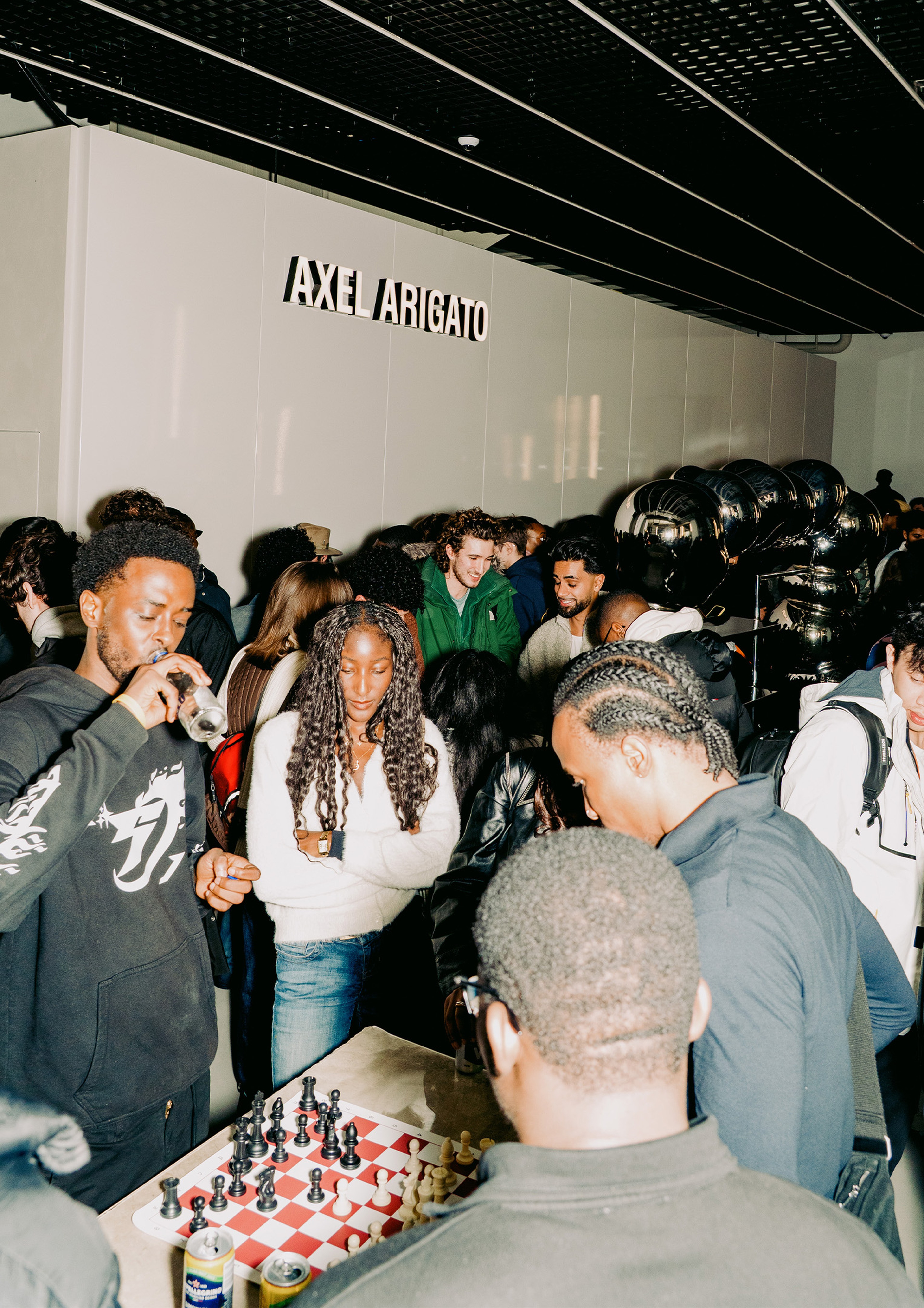 People gathered around a chessboard in a busy room with Axel Arigato signage, engaging in conversation and observing the game.