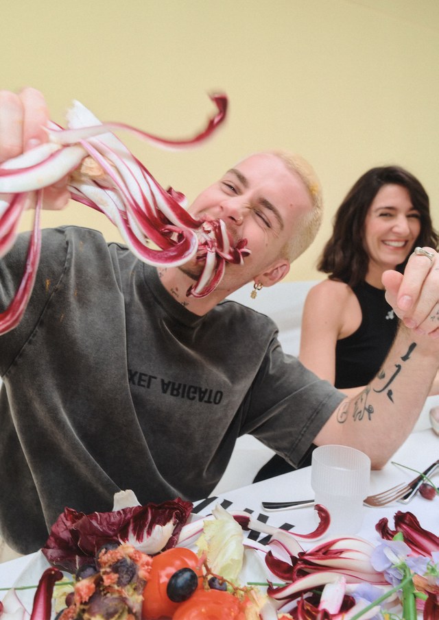Person playfully eating radicchio at a table, smiling, with another person laughing in the background. Plates of food are visible.