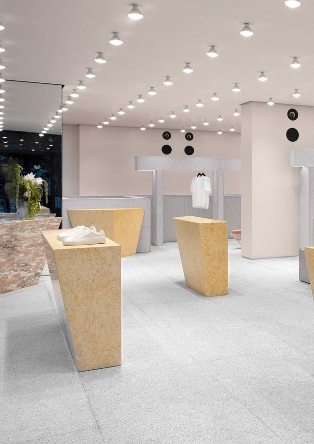 Modern retail store interior with beige and grey decor, geometric display tables holding white shoes and shirts, and bright overhead lighting.