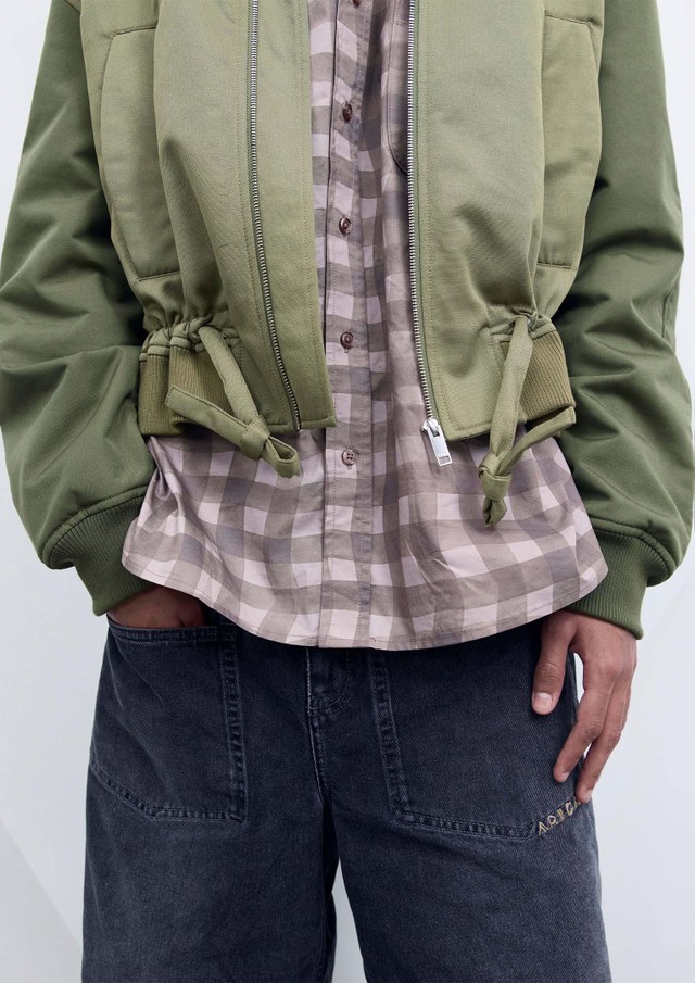 Person wearing a green jacket over a checked shirt with baggy dark jeans, standing against a light background.