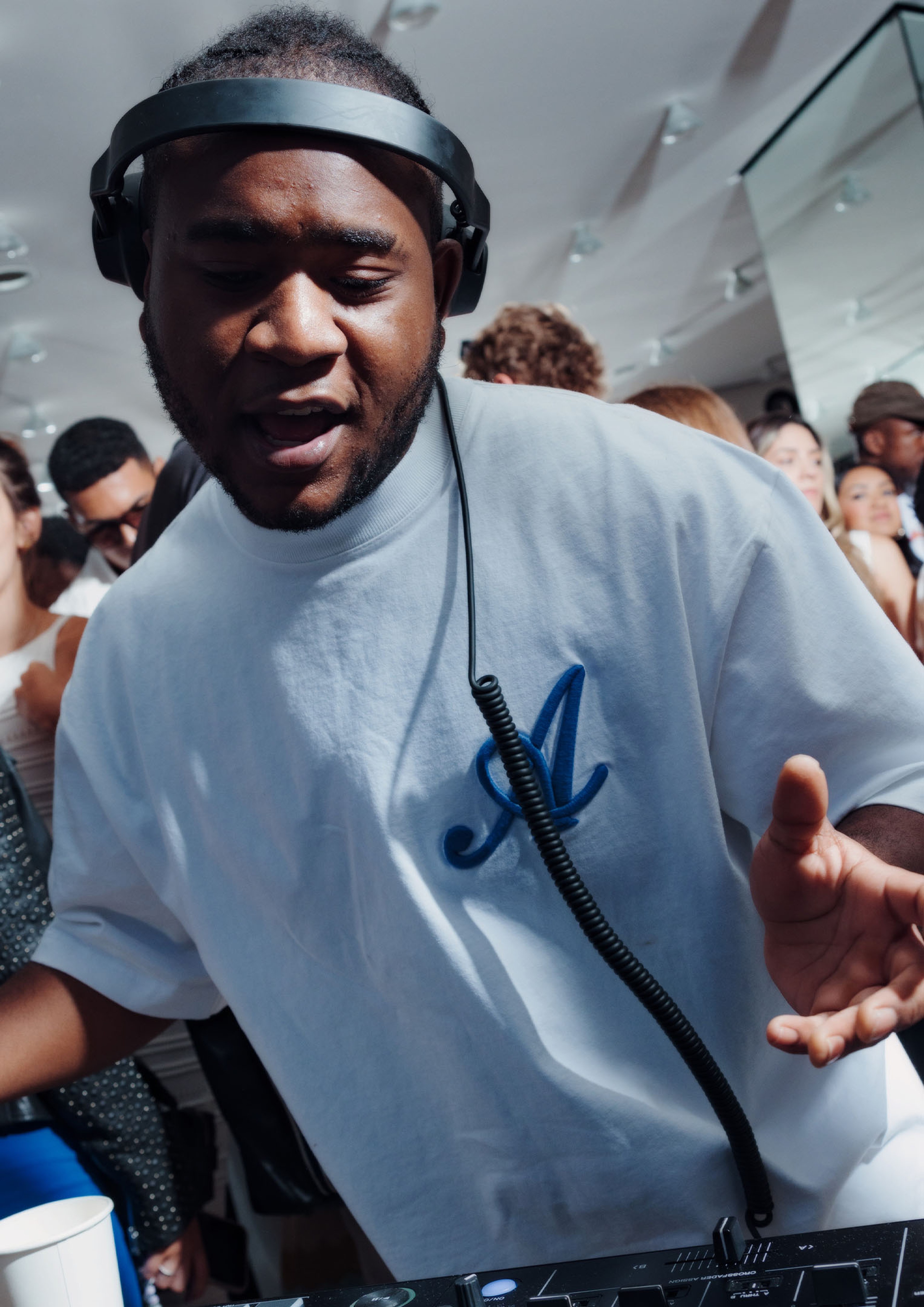 DJ wearing headphones and a white shirt with a blue "A" logo, performing at a lively indoor event with a crowd in the background.