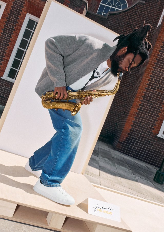 Person in a gray sweater and blue jeans playing a saxophone, standing on a platform with a brick building in the background.
