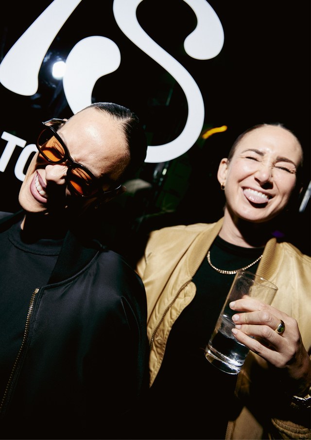 Two people smiling and posing playfully; one wearing sunglasses, the other holding a drink and sticking out their tongue.