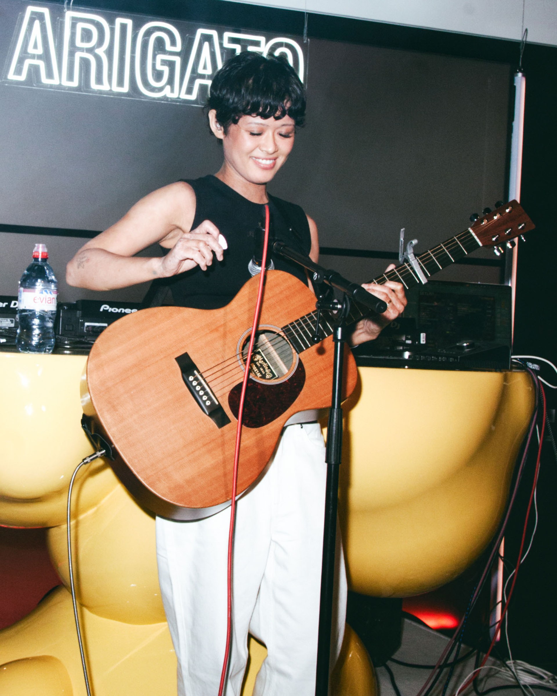 A person smiling while playing an acoustic guitar on stage, with a neon sign and sound equipment in the background.