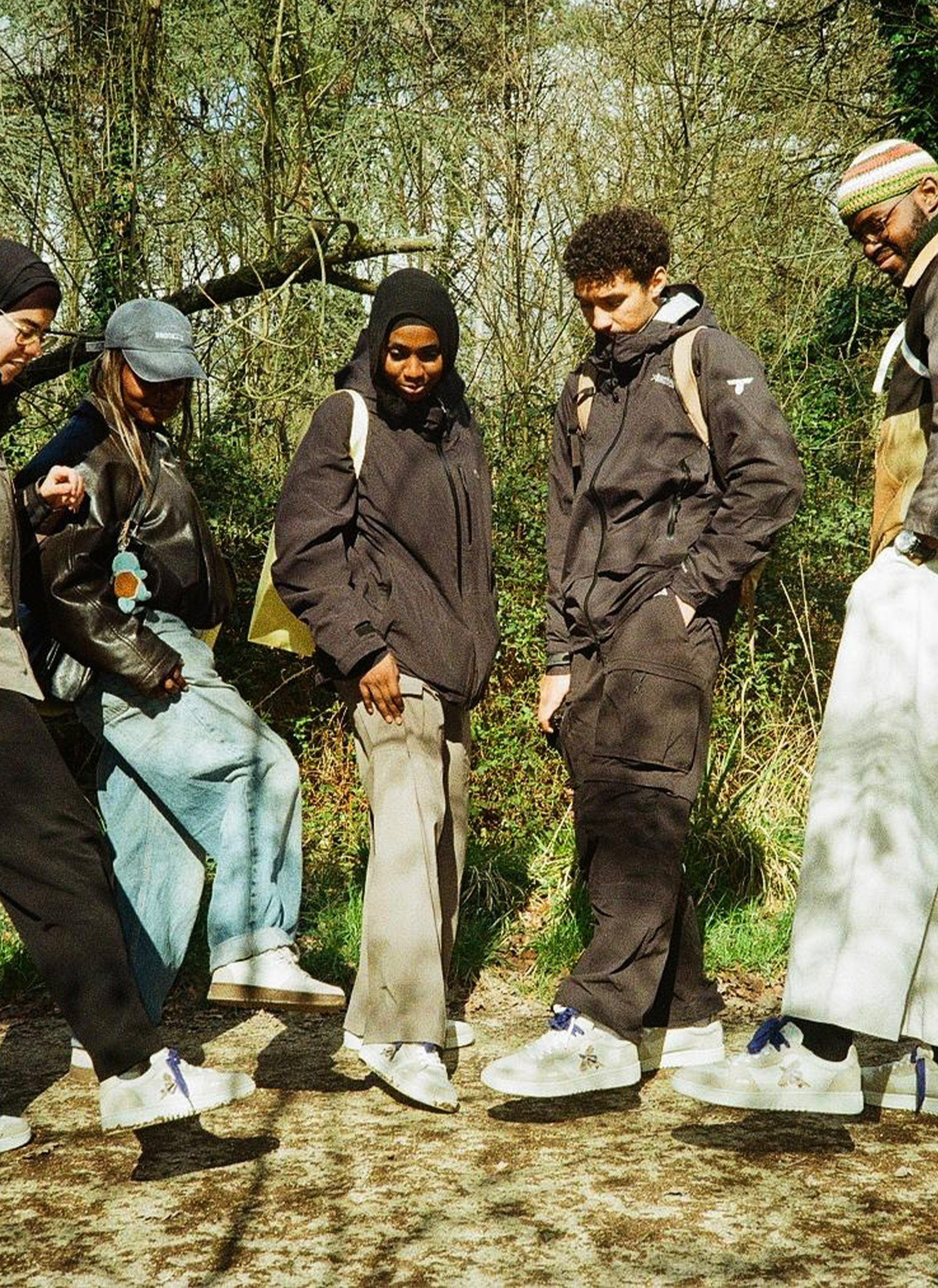 A group of five people in casual outdoor attire stand together on a path surrounded by trees, all looking down at their shoes.