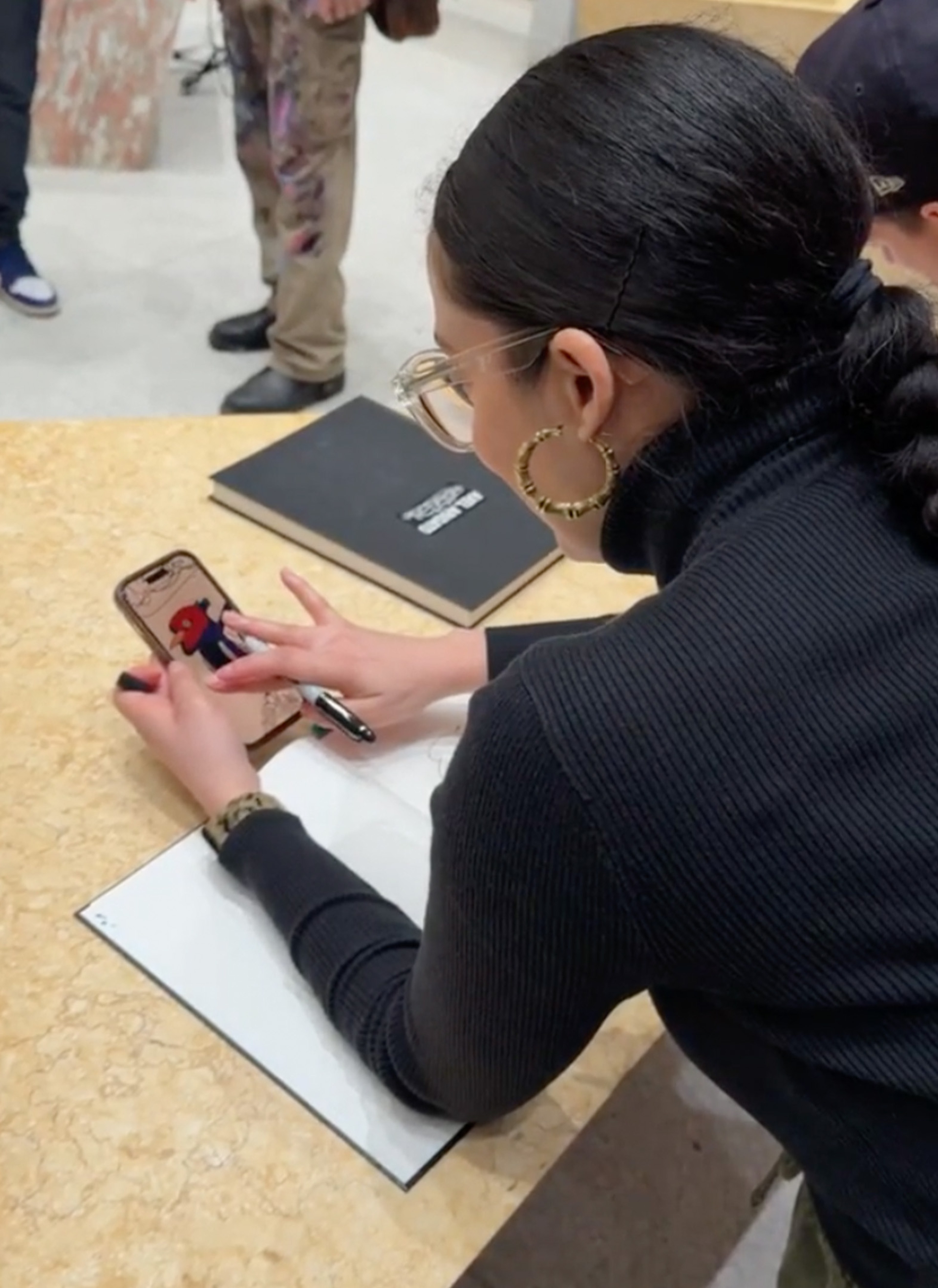 A woman in a black turtleneck looks at a phone while holding a pen, standing at a beige table with a closed notebook.