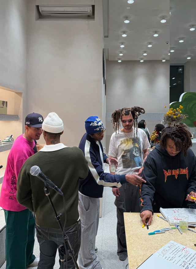 Five people gather around a table covered with papers and art supplies in a modern, well-lit room. One person is pointing at the table.