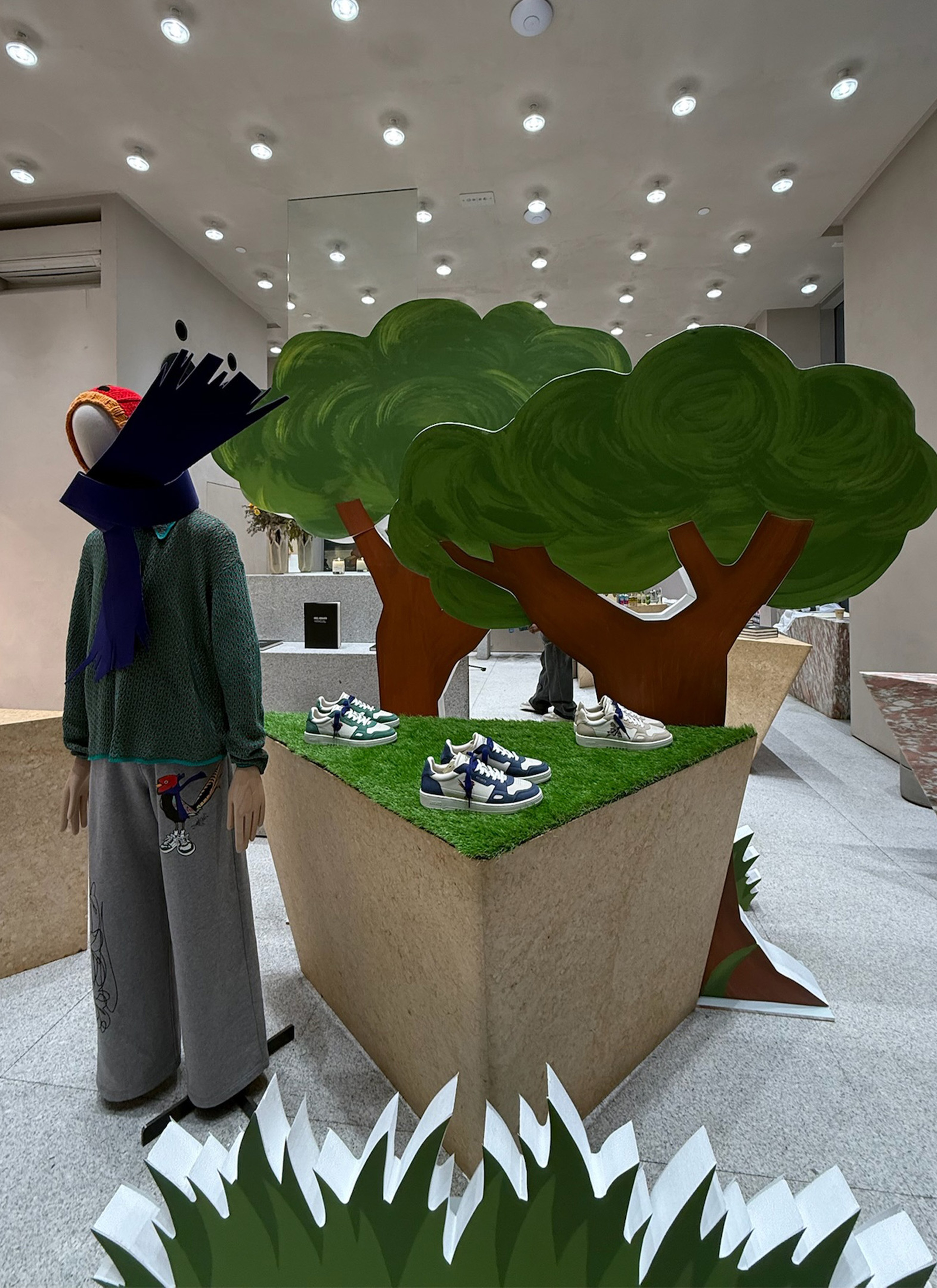Store display with mannequin in winter attire beside tree cutouts. Sneakers showcased on elevated platforms with grassy tops.