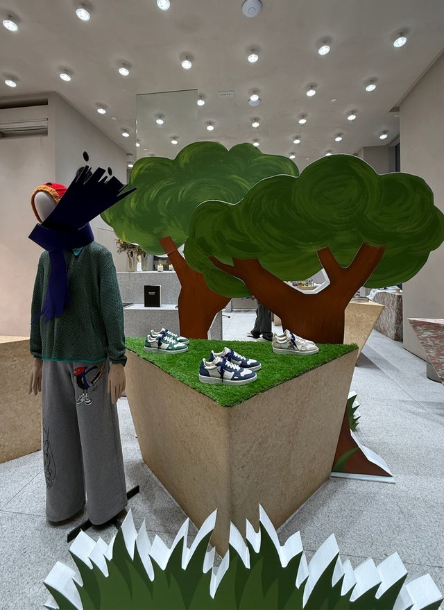 Store display with mannequin in winter attire beside tree cutouts. Sneakers showcased on elevated platforms with grassy tops.