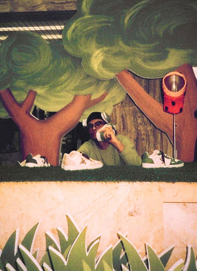 Man wearing sunglasses and headphones drinks from a bottle behind a display of sneakers, with tree decorations in the background.