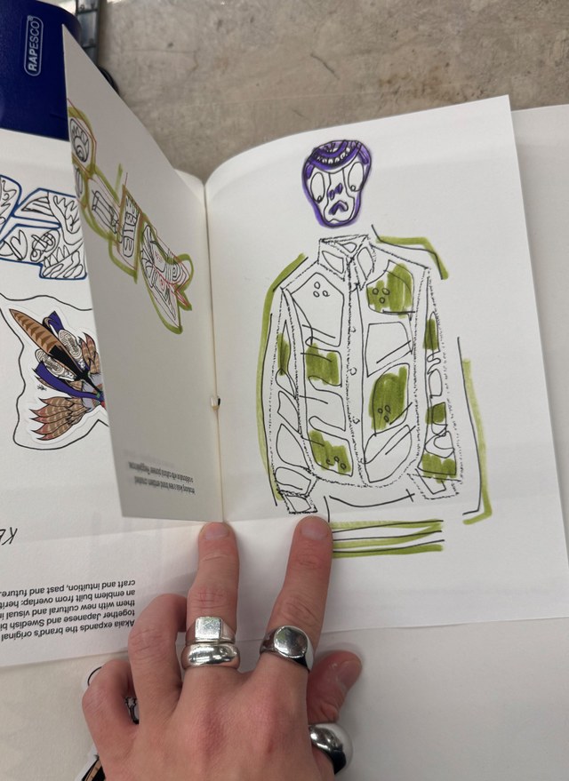Hand with rings holding open sketchbook showing an abstract drawing of a person in patterned attire with green and purple accents.