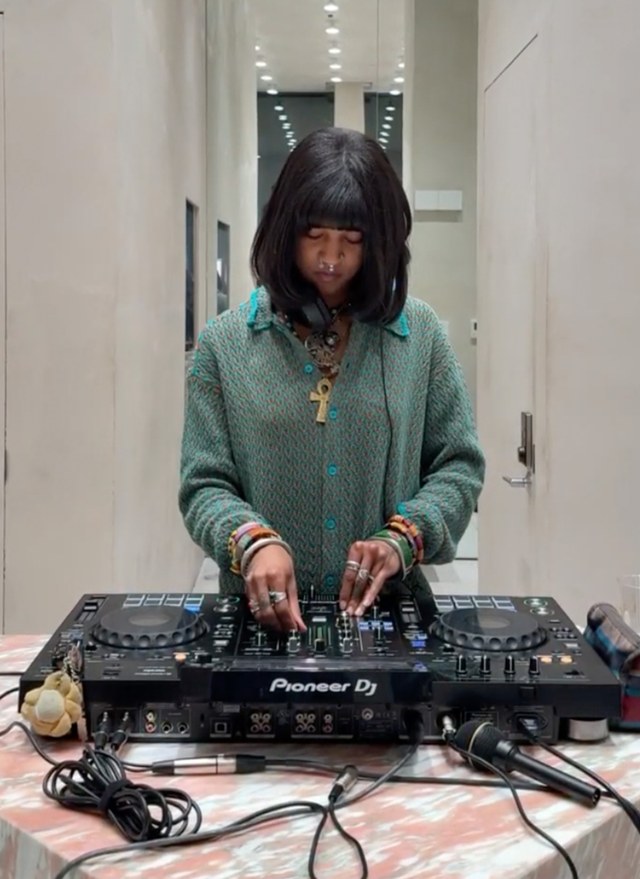 DJ at a mixer deck, wearing a green sweater and headphones, standing in a modern room with a marble table.