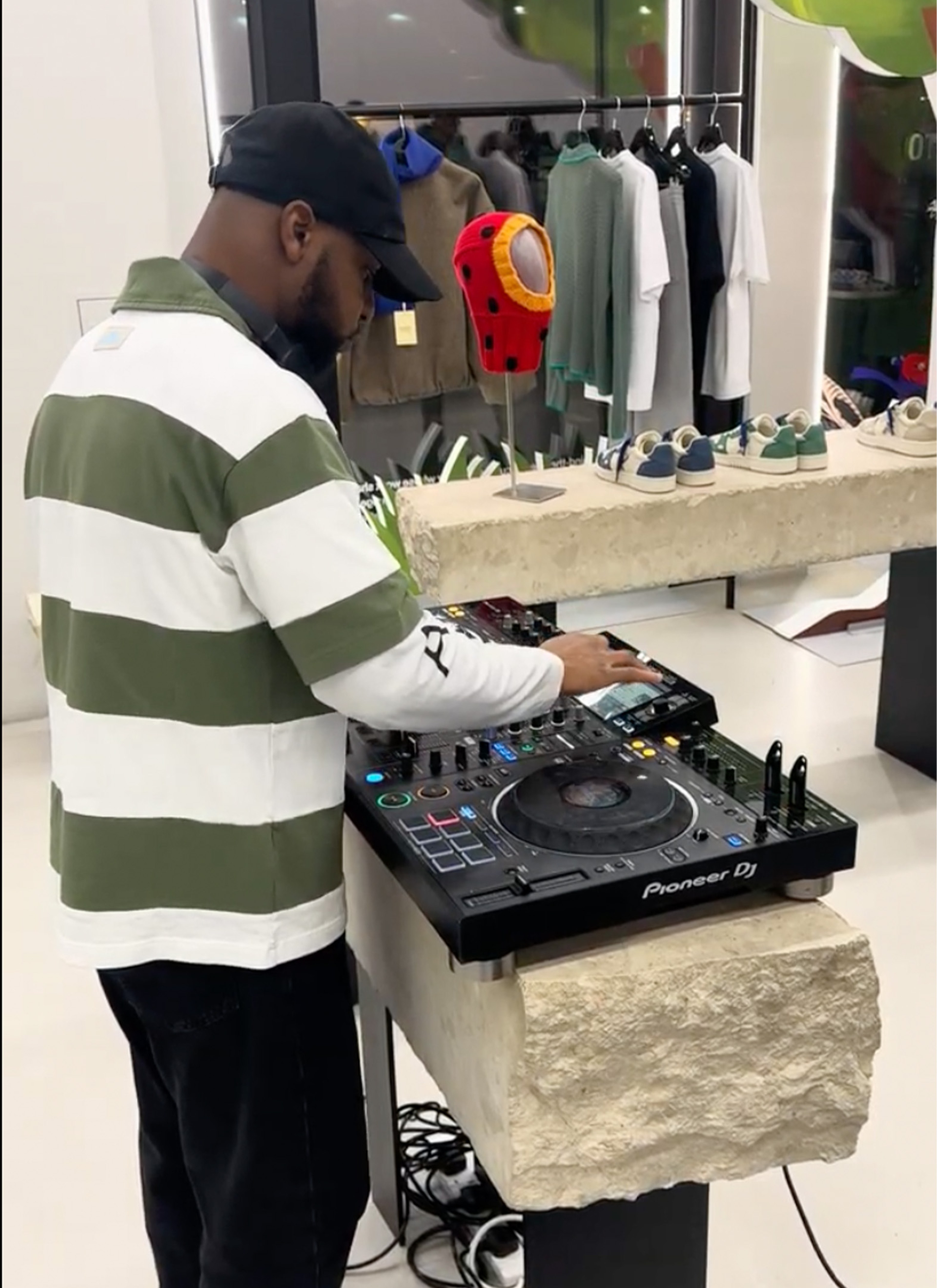 DJ wearing a cap and striped shirt mixing music on Pioneer DJ equipment in a modern clothing store.