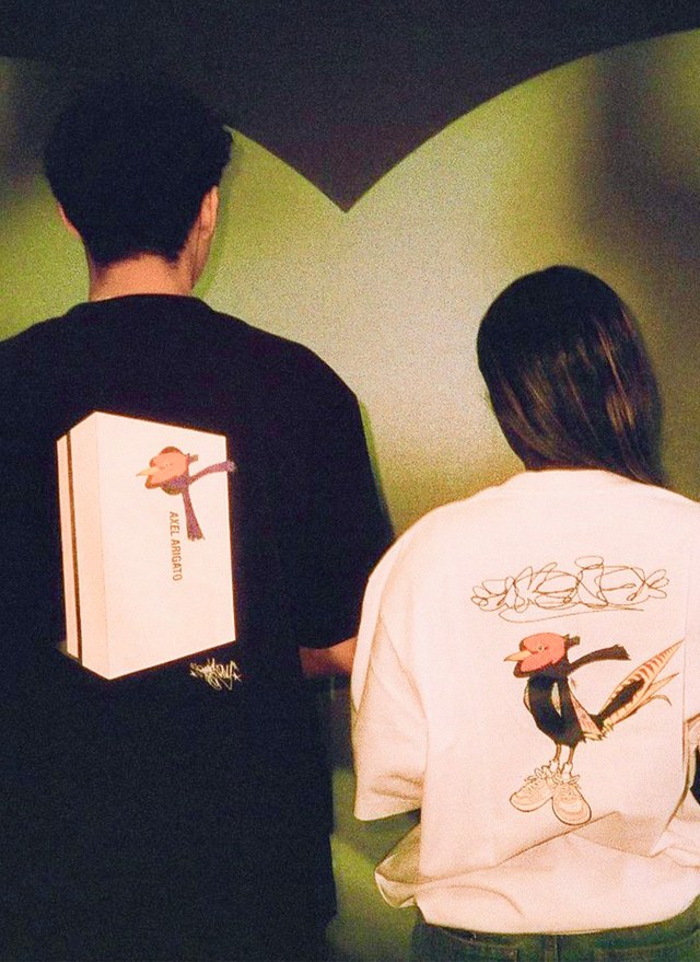 Two people stand with their backs to the camera, wearing shirts featuring cartoon-like characters in sneakers on the back.