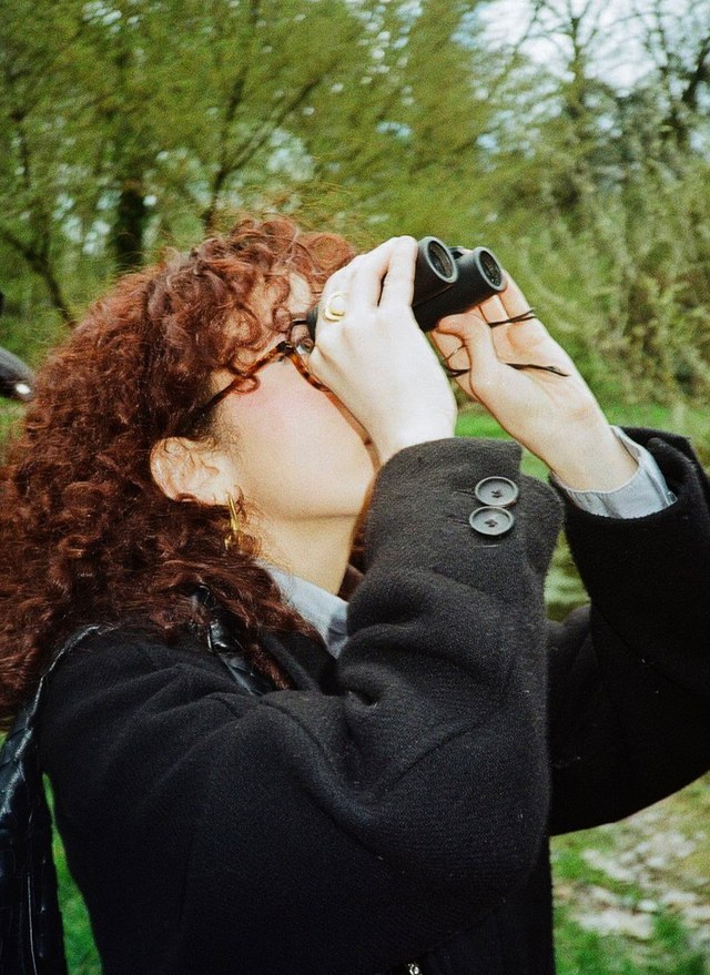 Person with curly hair wearing a black coat looking through binoculars in a wooded area.