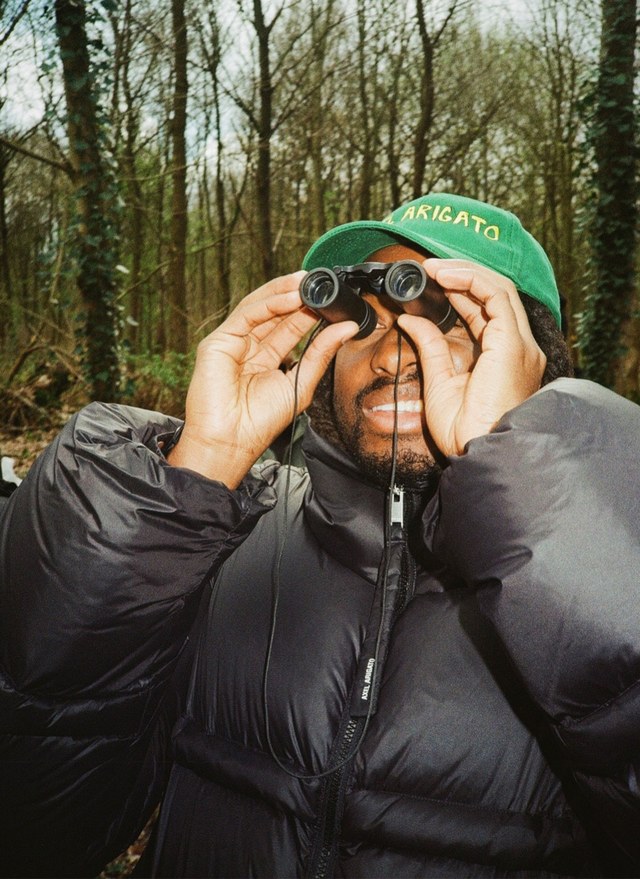 Person in a puffy jacket and green cap using binoculars in a wooded area.