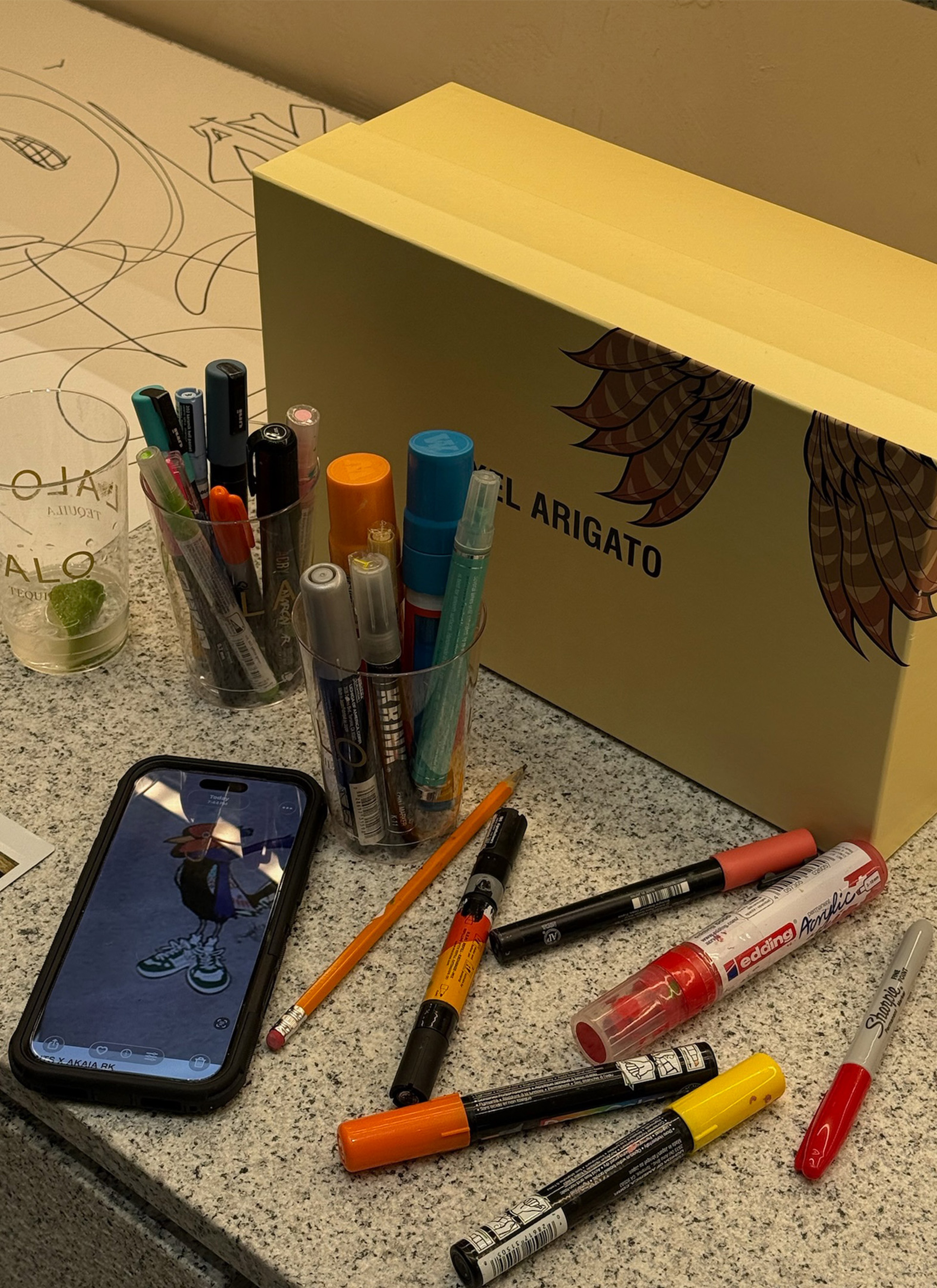 A desk with markers, pens, a mobile phone displaying an illustration, and a yellow box with wing artwork and "Arigato" text.