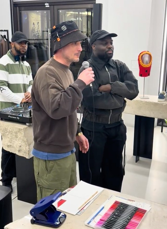 Three people indoors; one speaks into a microphone, wearing a bucket hat. Another stands beside him, and the third is at a DJ booth.