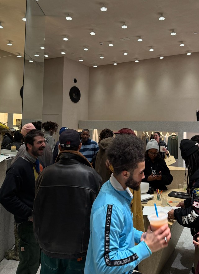 People gathered in a modern store, chatting and enjoying drinks under bright ceiling lights. Mirrors reflect part of the group.
