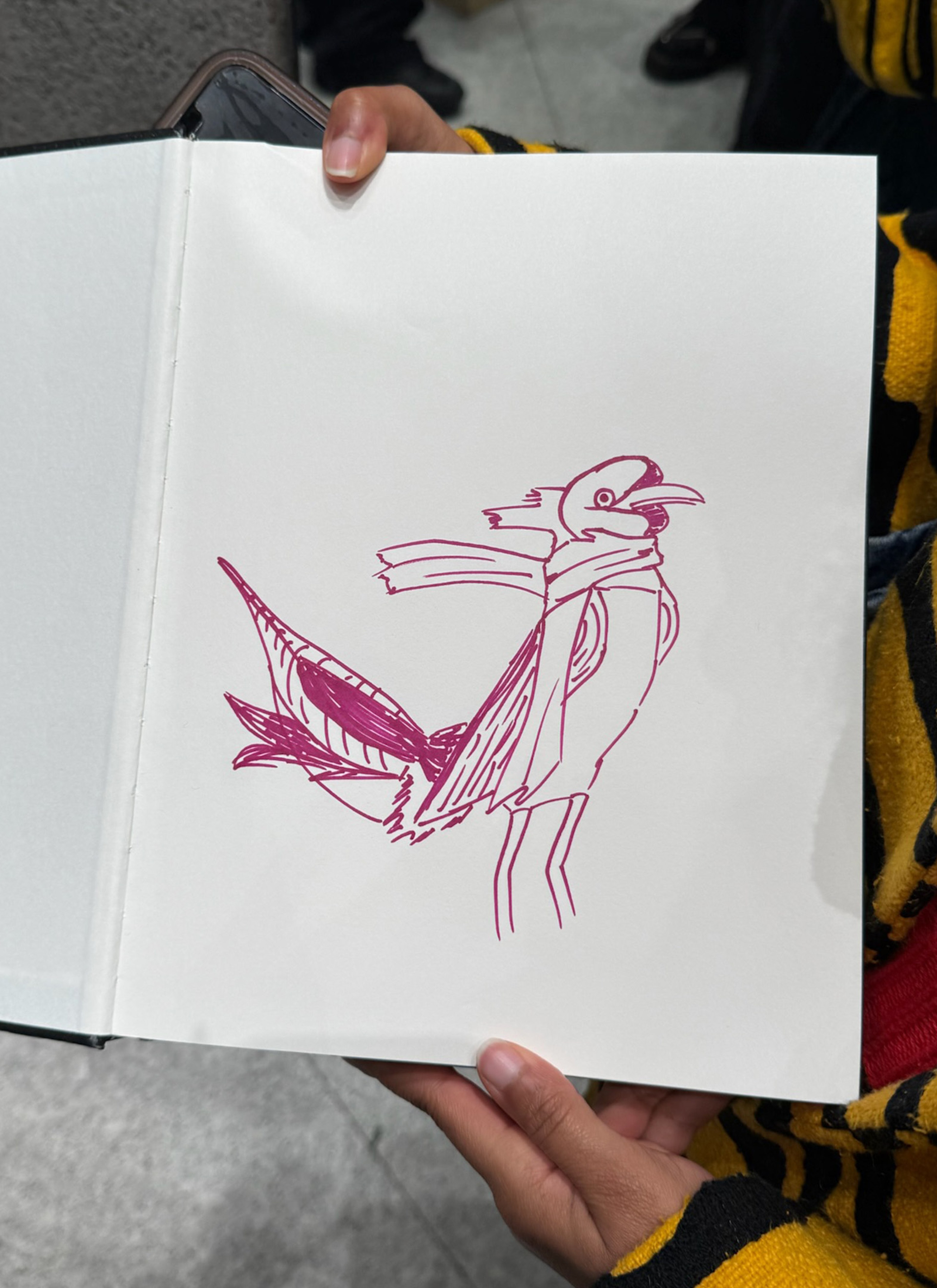 Person holding an open sketchbook displaying a simple, abstract drawing of a bird using red lines.