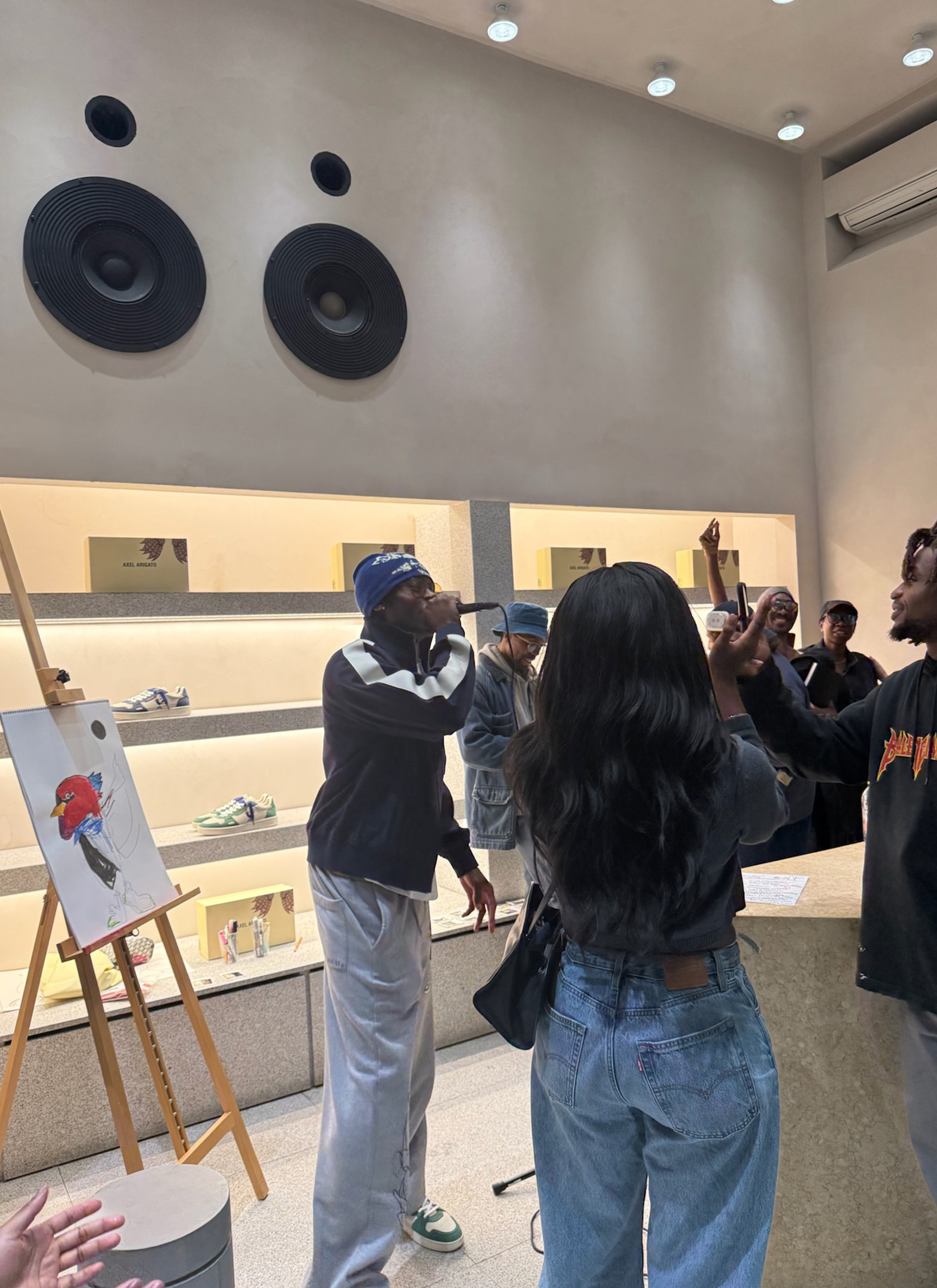 A hip-hop artist performs to a small crowd in a modern retail store, with shoes displayed in the background and a painting on an easel nearby.