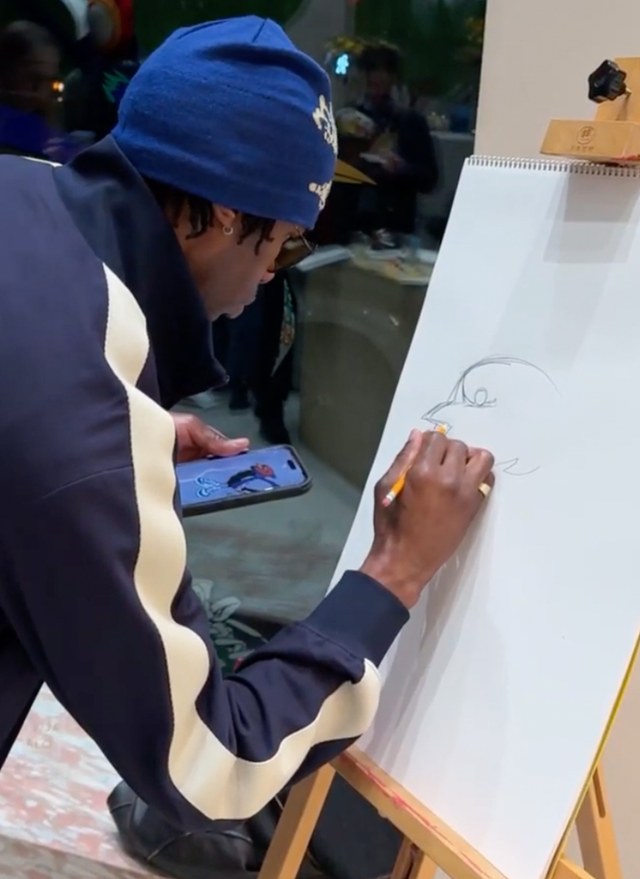 Person in a blue hat drawing on a sketchpad using a pencil, with a phone showing a reference image in the other hand.