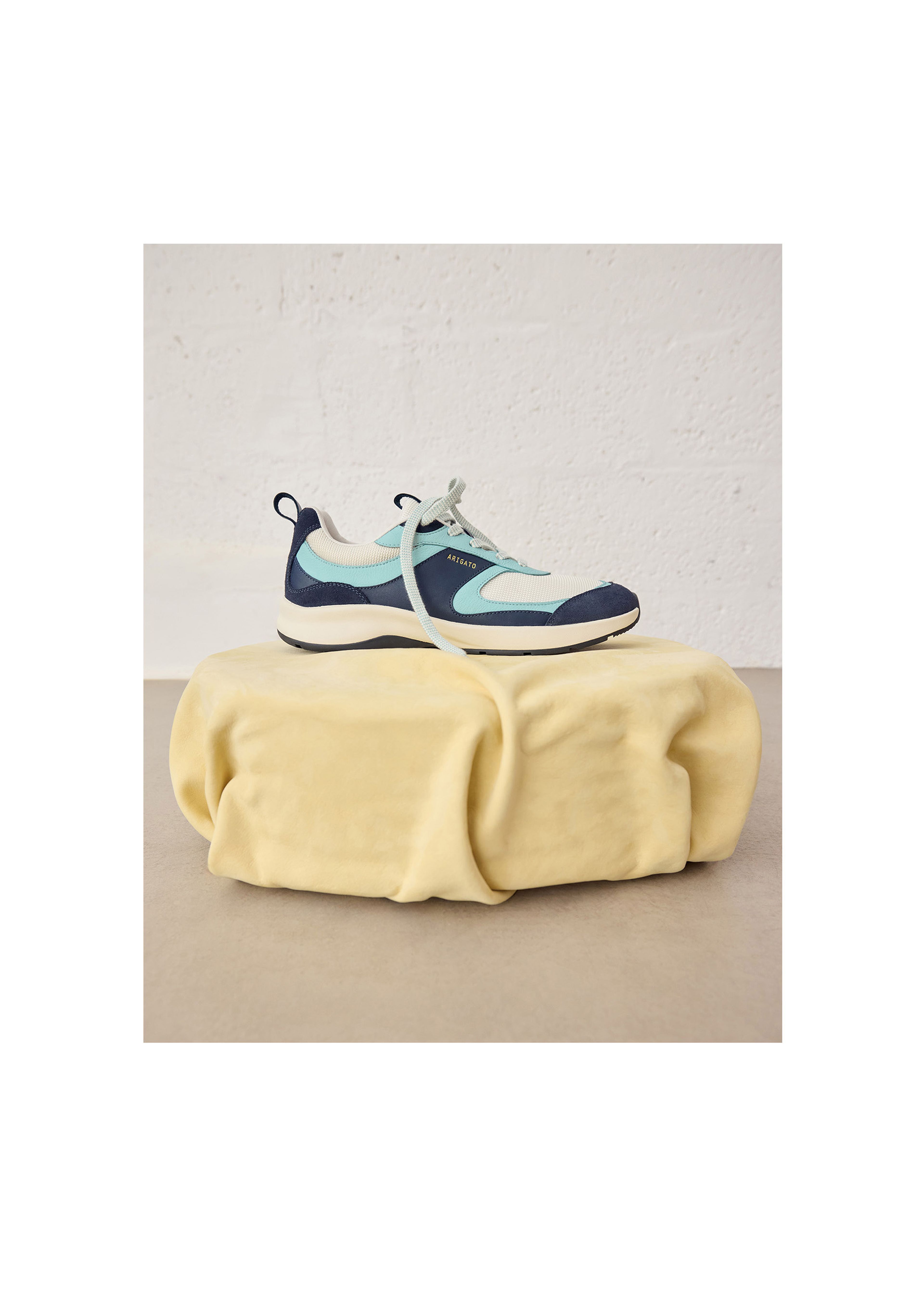 A light blue and navy sneaker with a white sole rests on a soft, round, yellow satin cushion against a textured white wall.