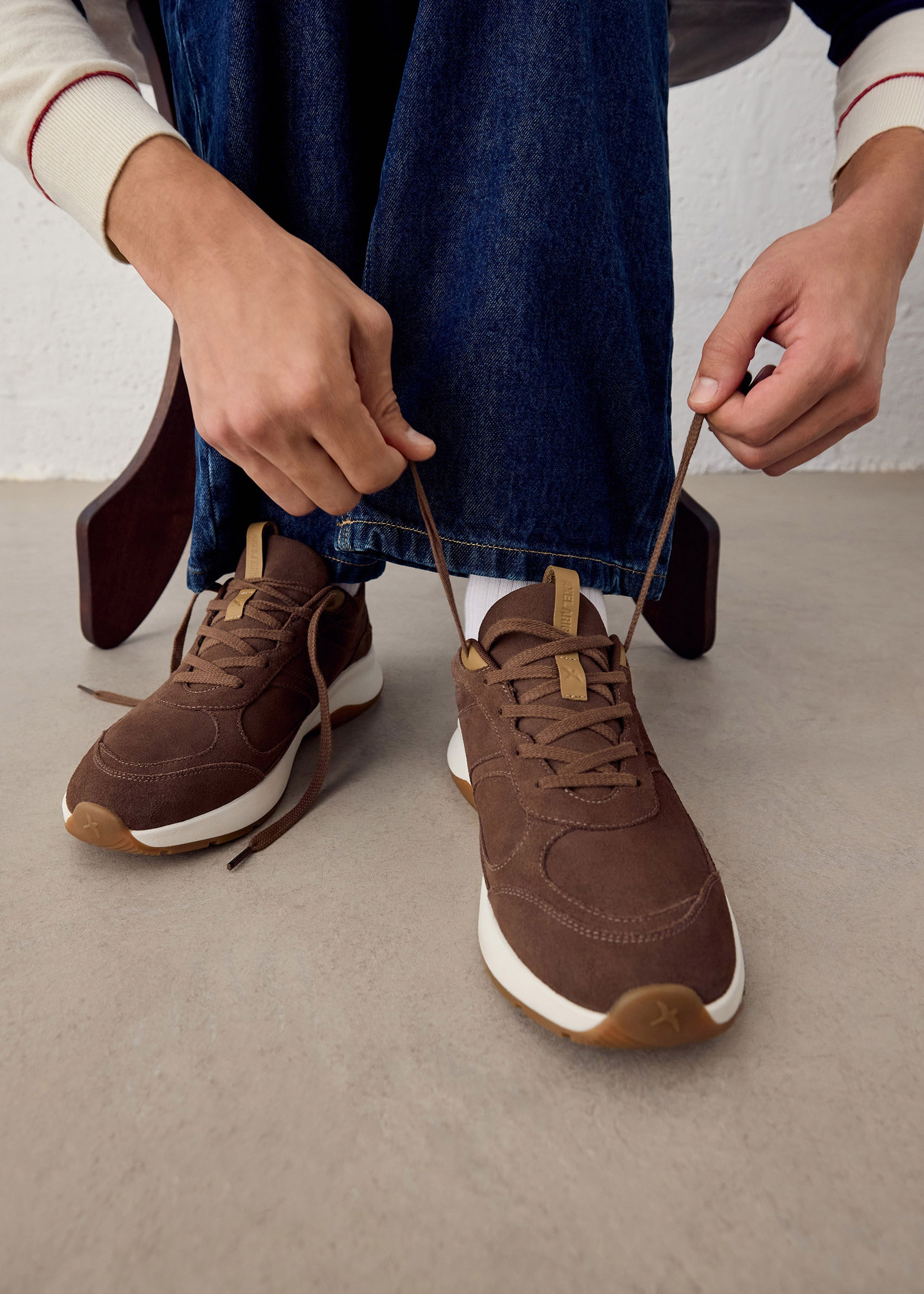 Person tying brown sneakers on a light brown floor, wearing blue jeans and a long-sleeve shirt.