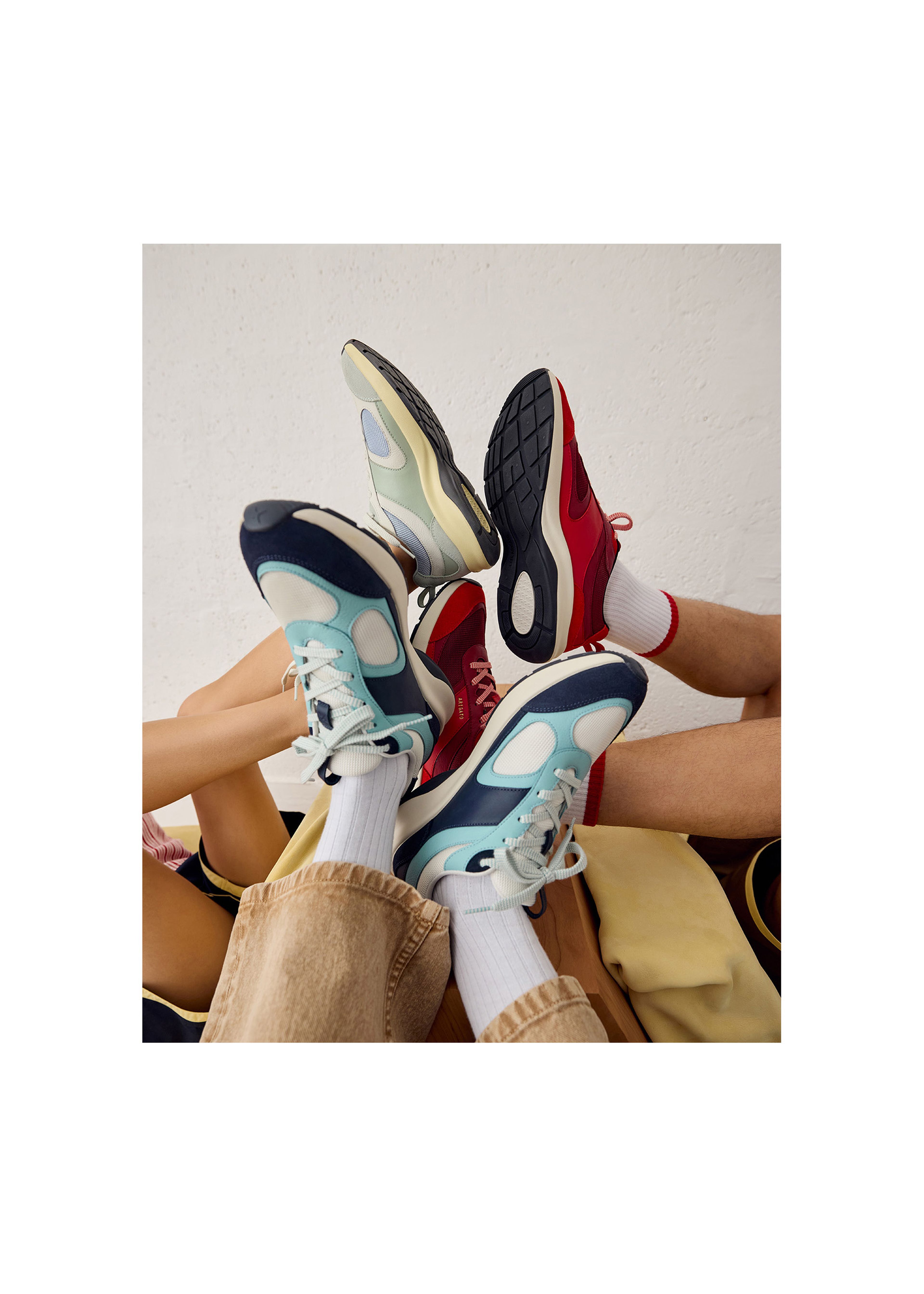 Four pairs of colorful sneakers, including blue, red, and beige, are playfully intertwined in the air against a white background.