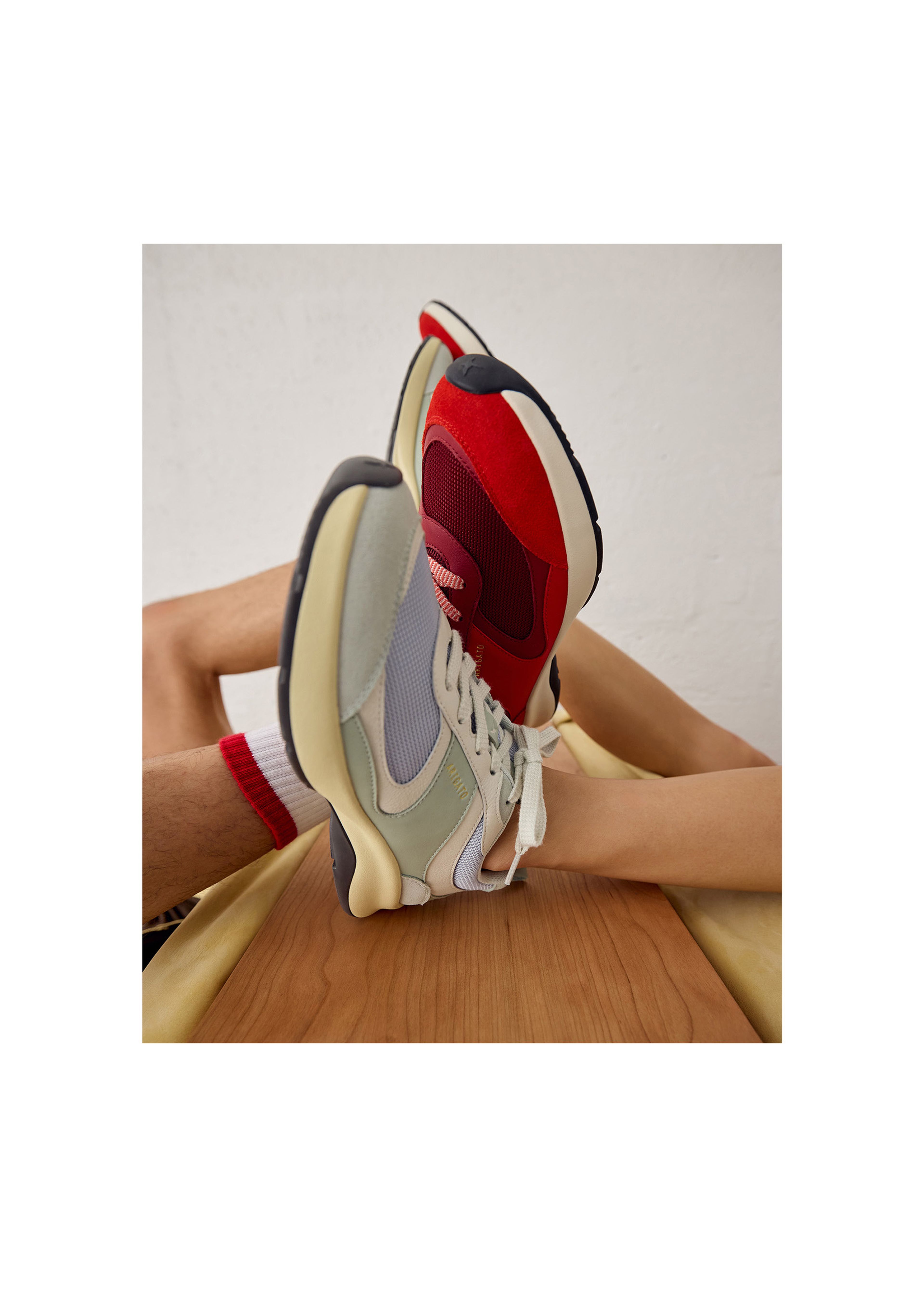 Two people wearing sneakers lie on their backs, feet up. One sneaker is red and black, the other is pale green and gray. A wooden surface is visible.