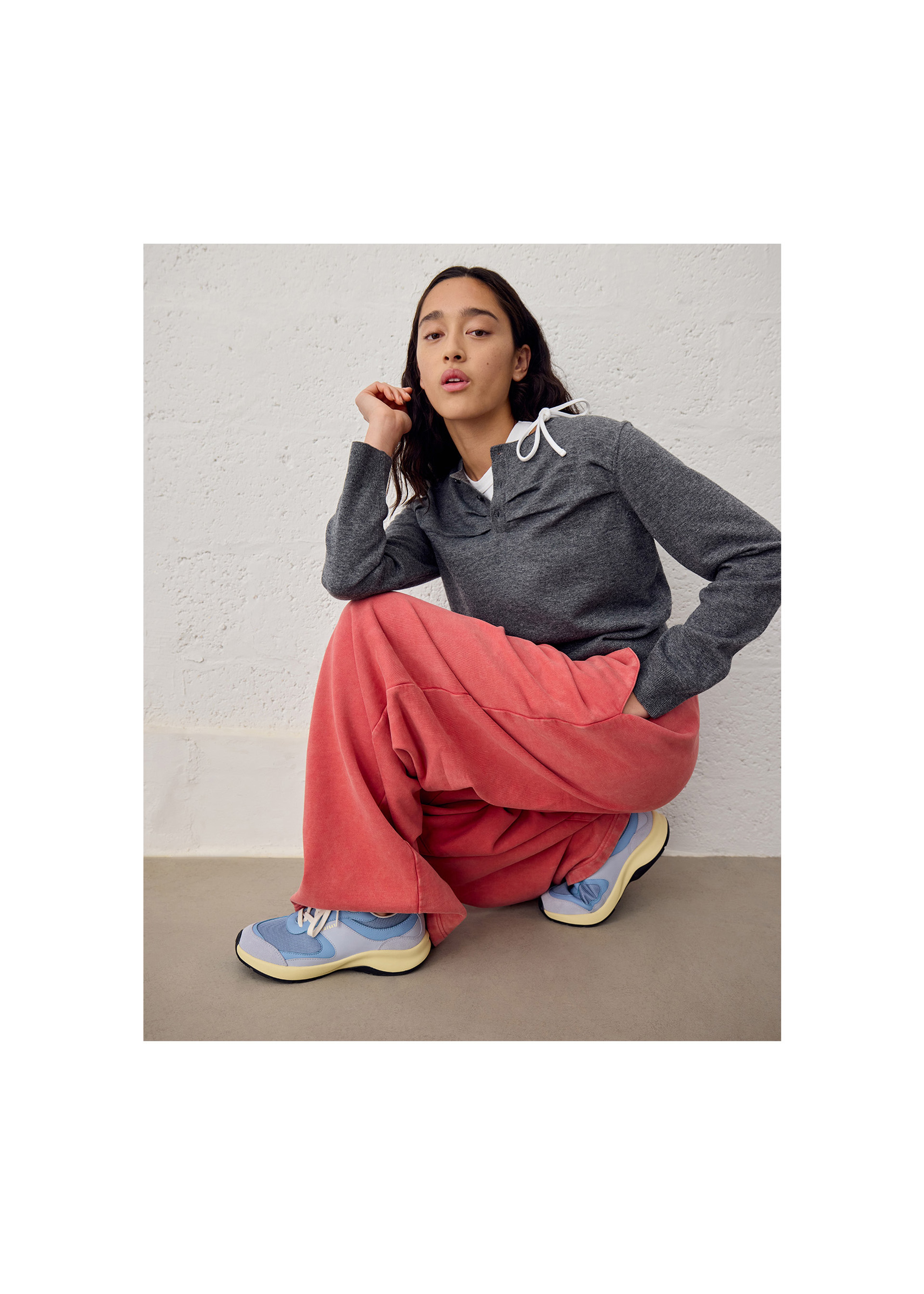 Person posing while crouching, wearing a gray hoodie, bright pink pants, and blue sneakers against a light textured background.