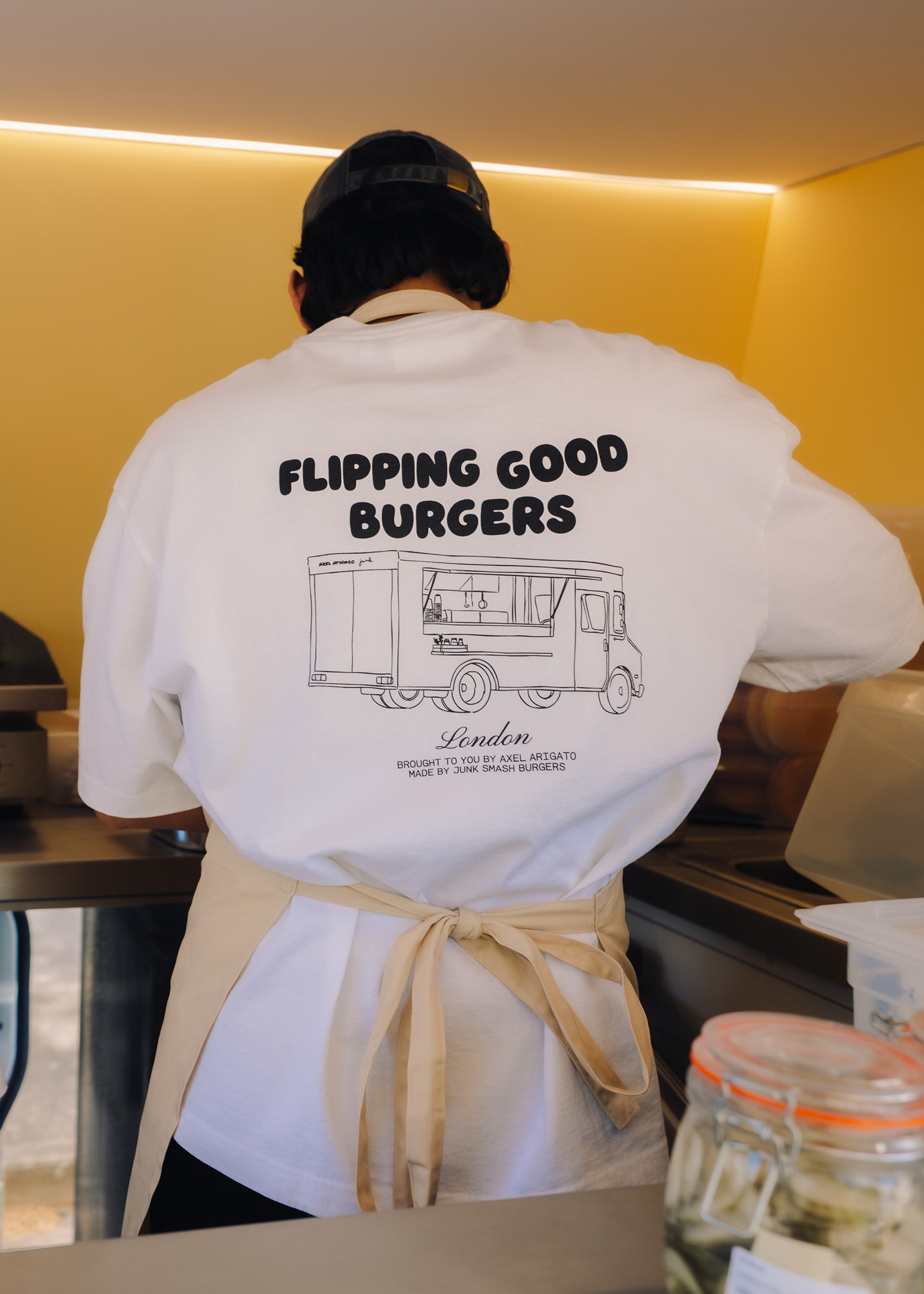 Person in a white T-shirt with "Flipping Good Burgers" text, working inside a food truck wearing an apron and cap, prepping food.