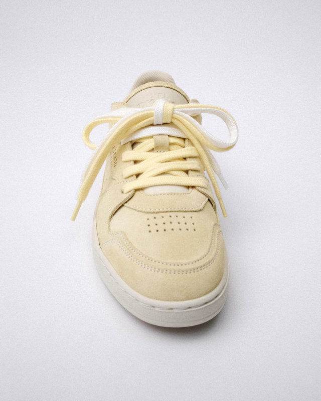 Cream-colored sneaker with matching laces and perforated details on the toe, viewed from the front against a plain background.