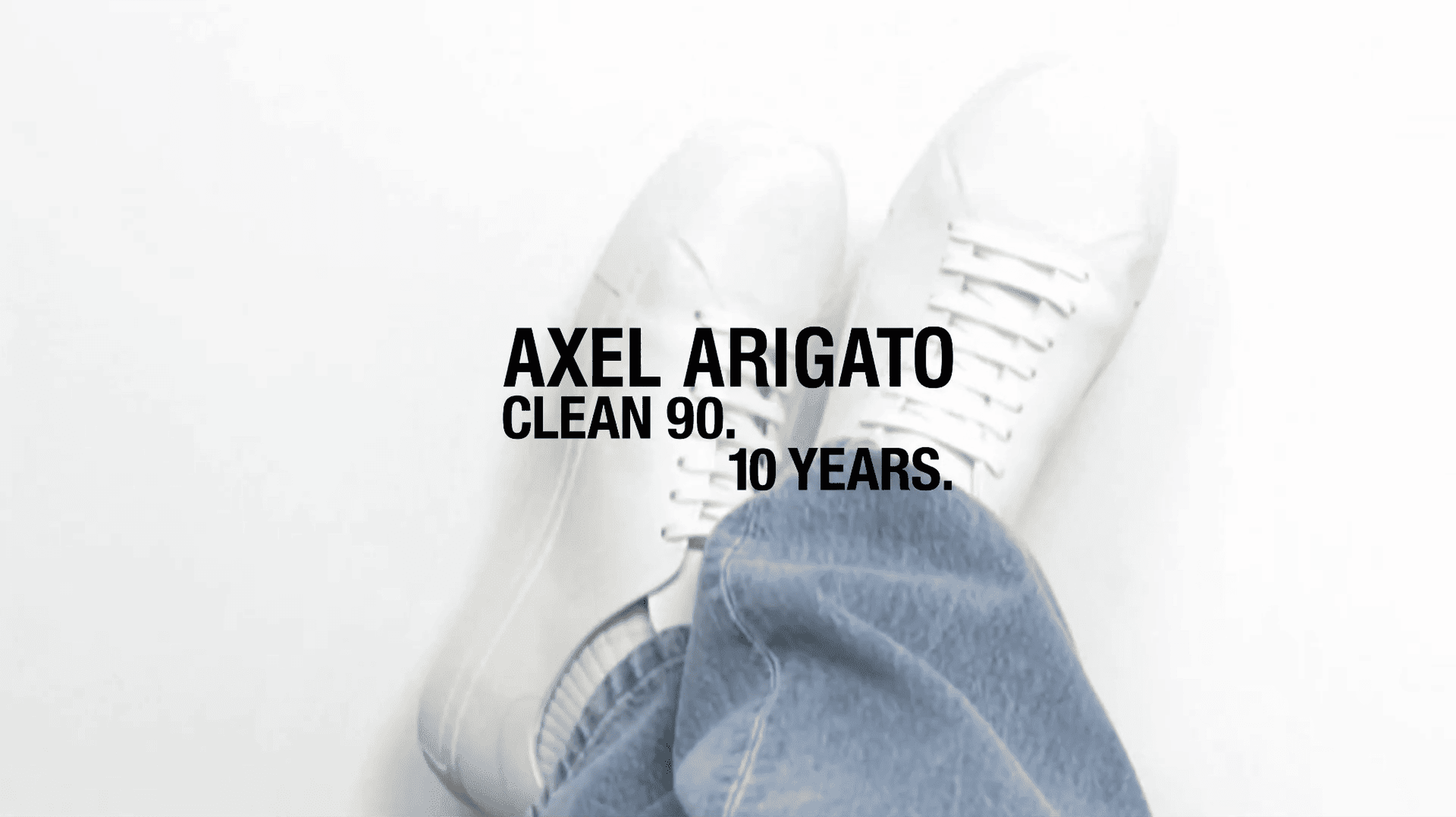 White sneakers labeled "Axel Arigato Clean 90. 10 Years" on a white background, partially covered by blue jeans.