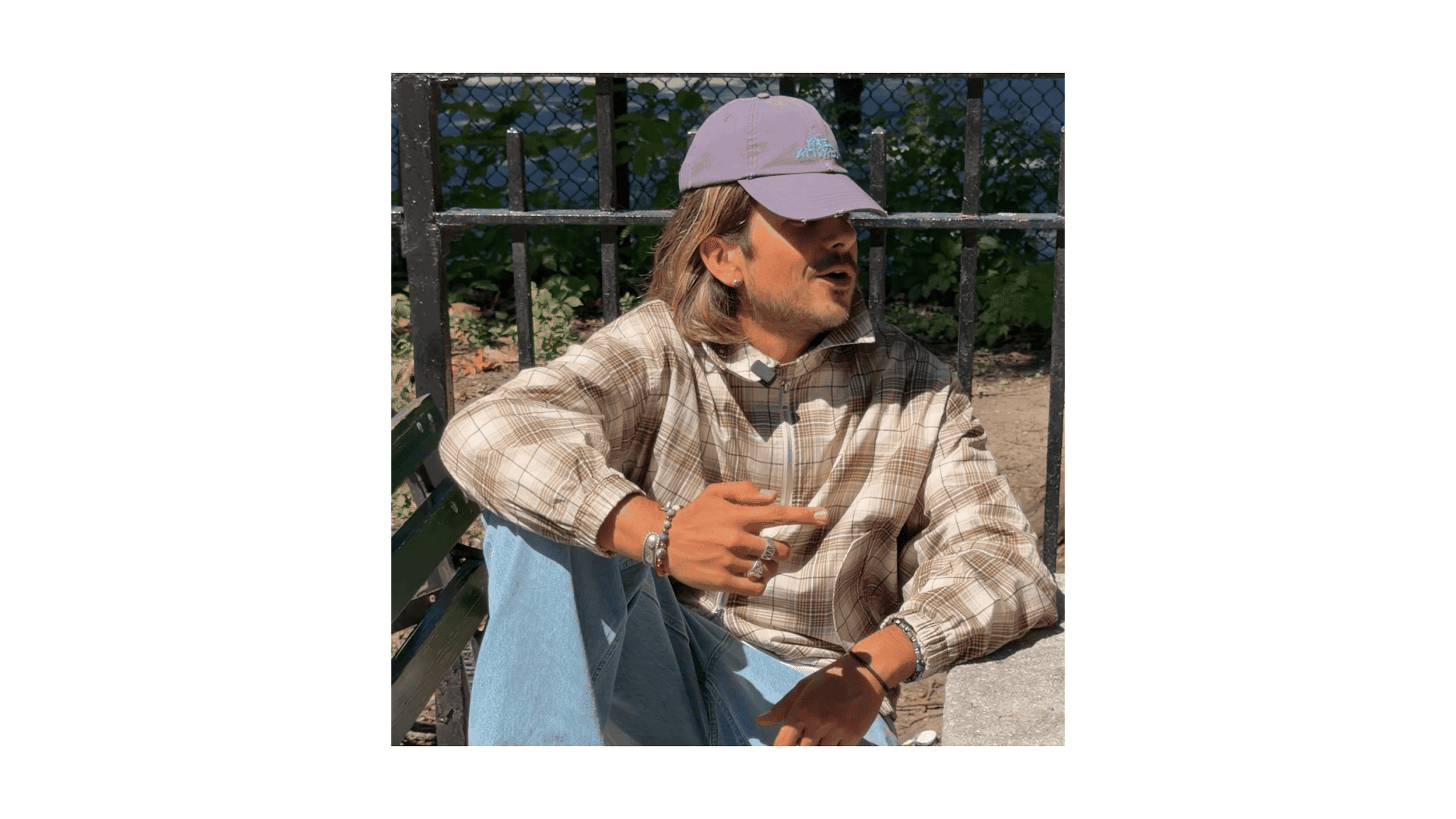 Man in a plaid shirt and cap sitting casually on a bench, framed by a metal fence and greenery in the background.