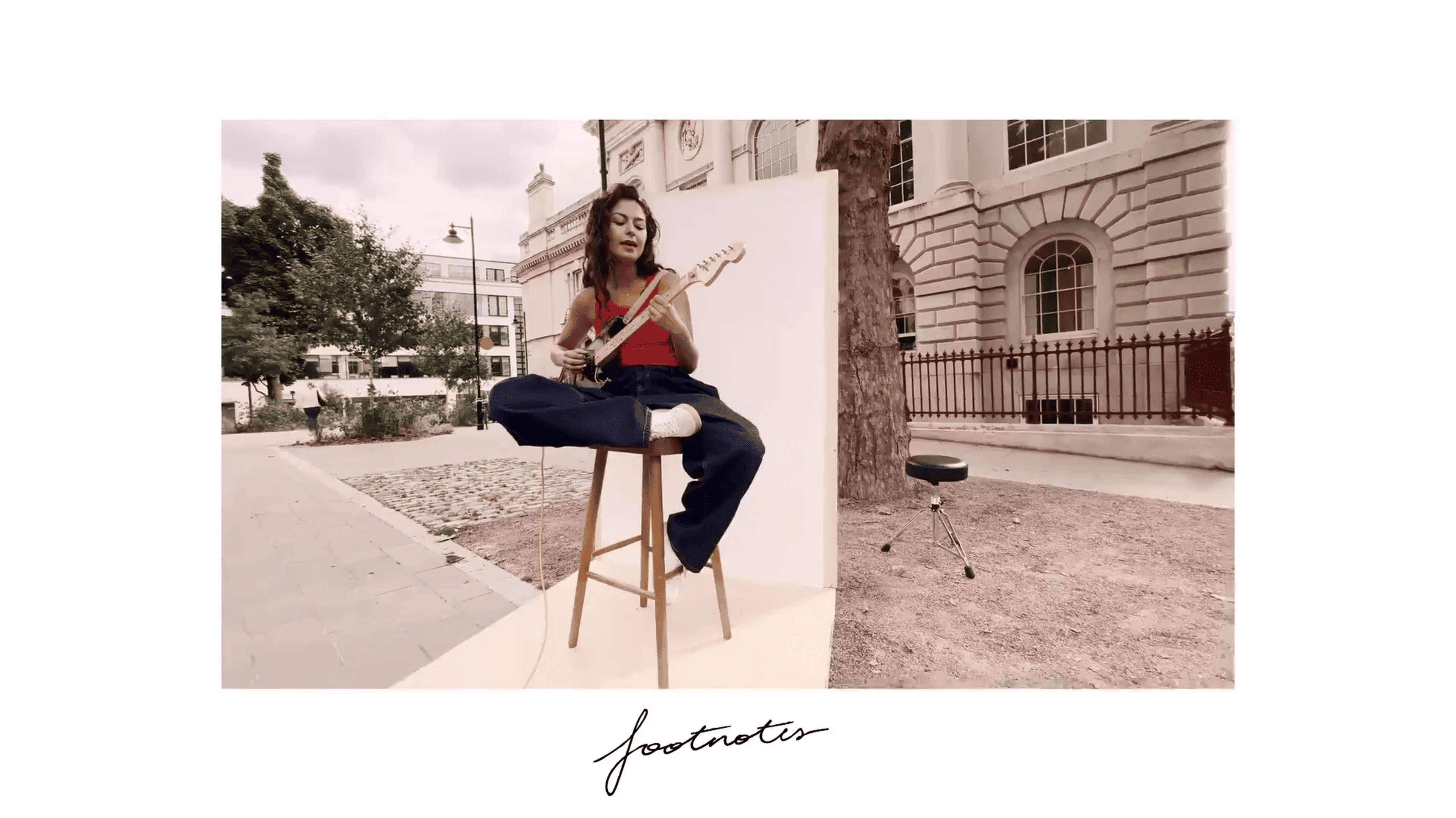 Woman playing guitar while sitting on a stool outdoors, with buildings and trees in the background. Text "footnote" at the bottom.