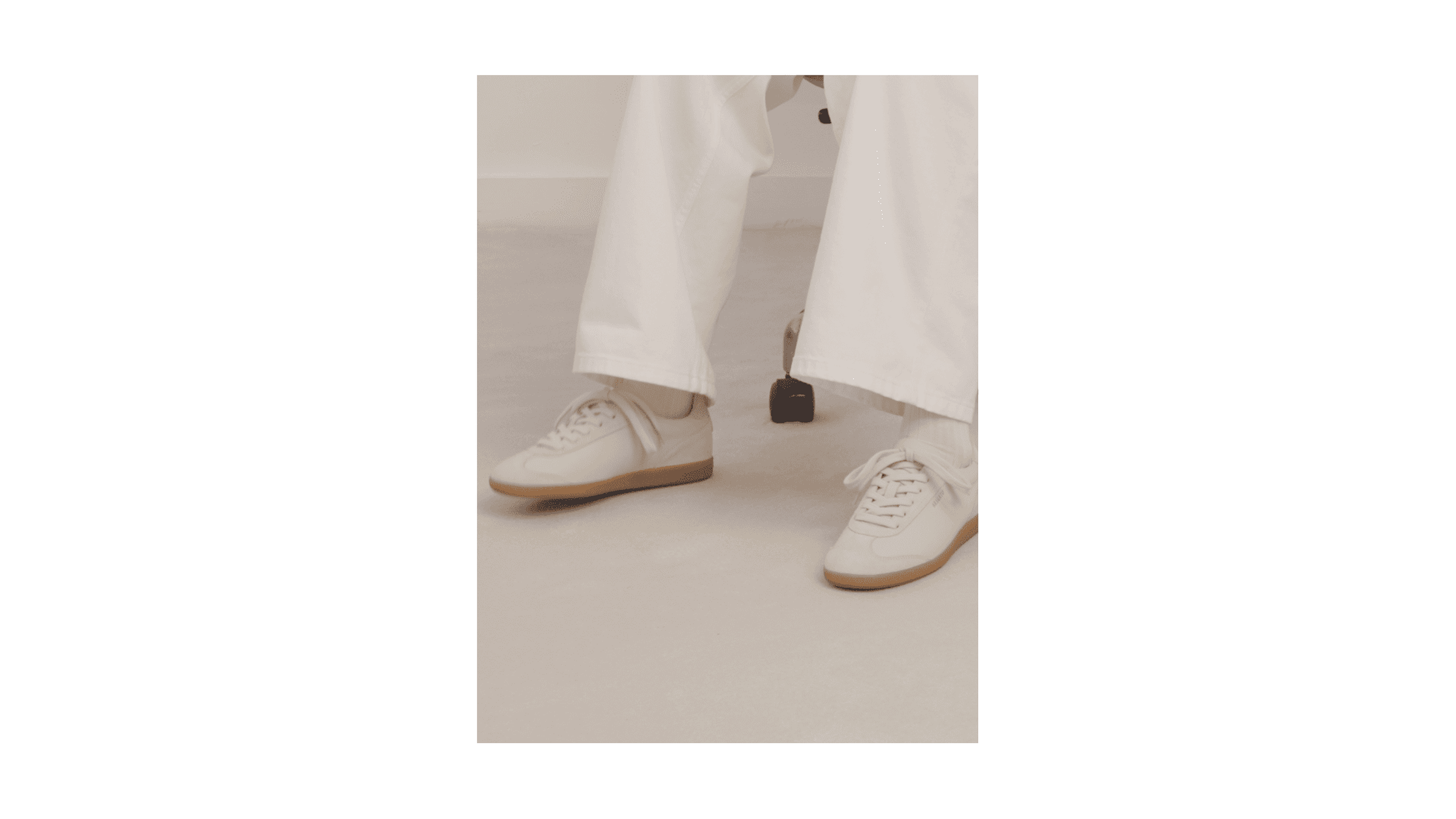 Person wearing white sneakers and white pants, seated on a chair, against a light-colored background.