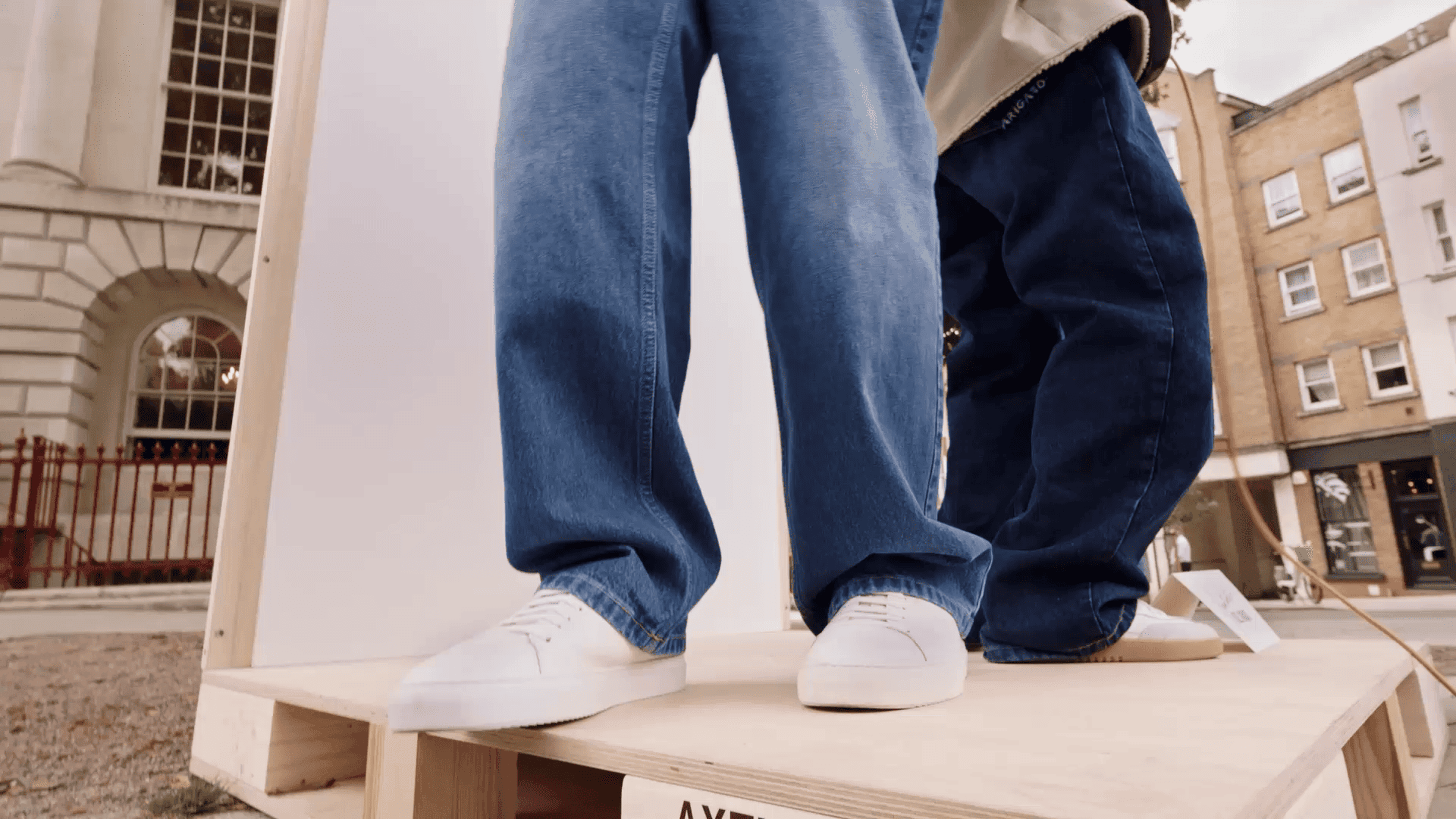 Low-angle view of two people wearing baggy jeans and white sneakers, standing on a wooden platform in an urban outdoor setting.